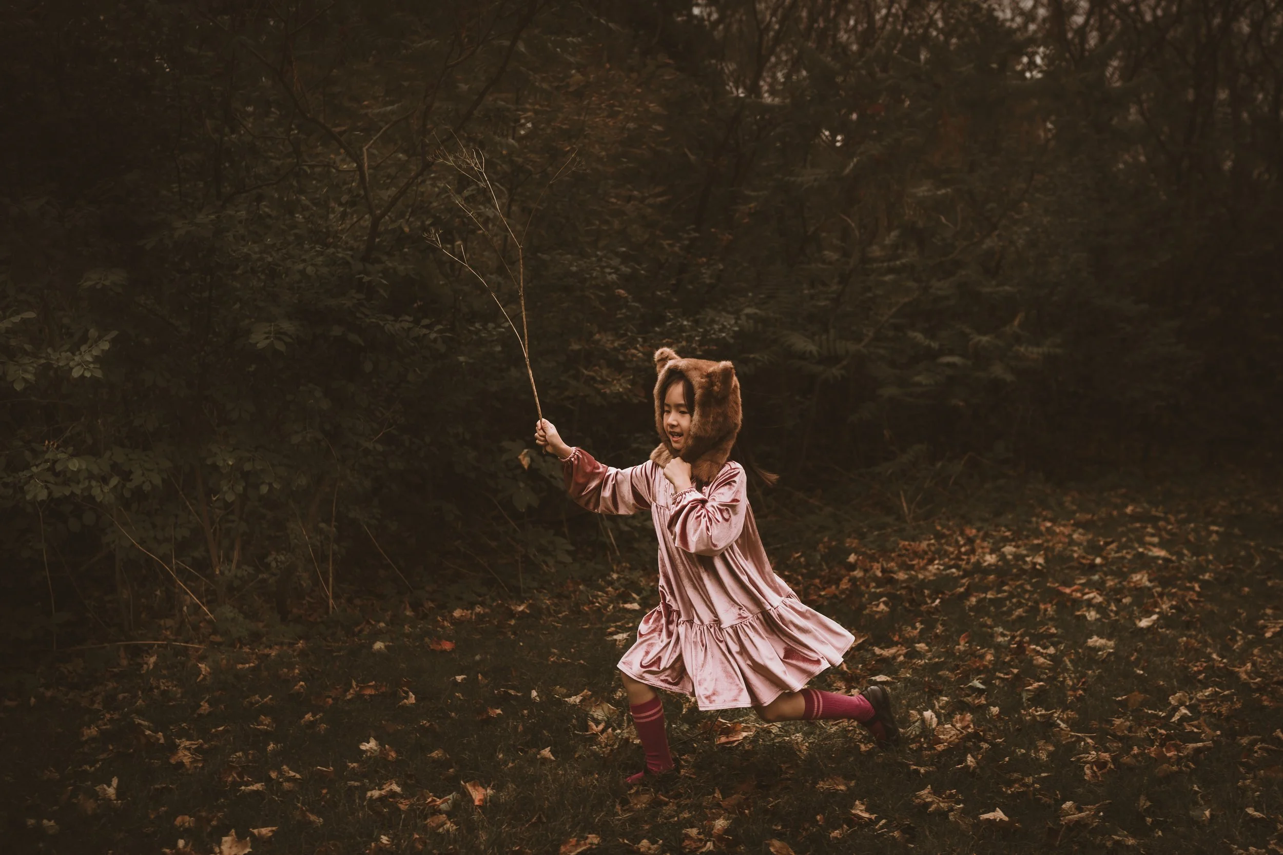 A young girl dressed in a pink dress and striped socks running through a wooded area during fall, wearing a bear costume hat and holding a stick.
