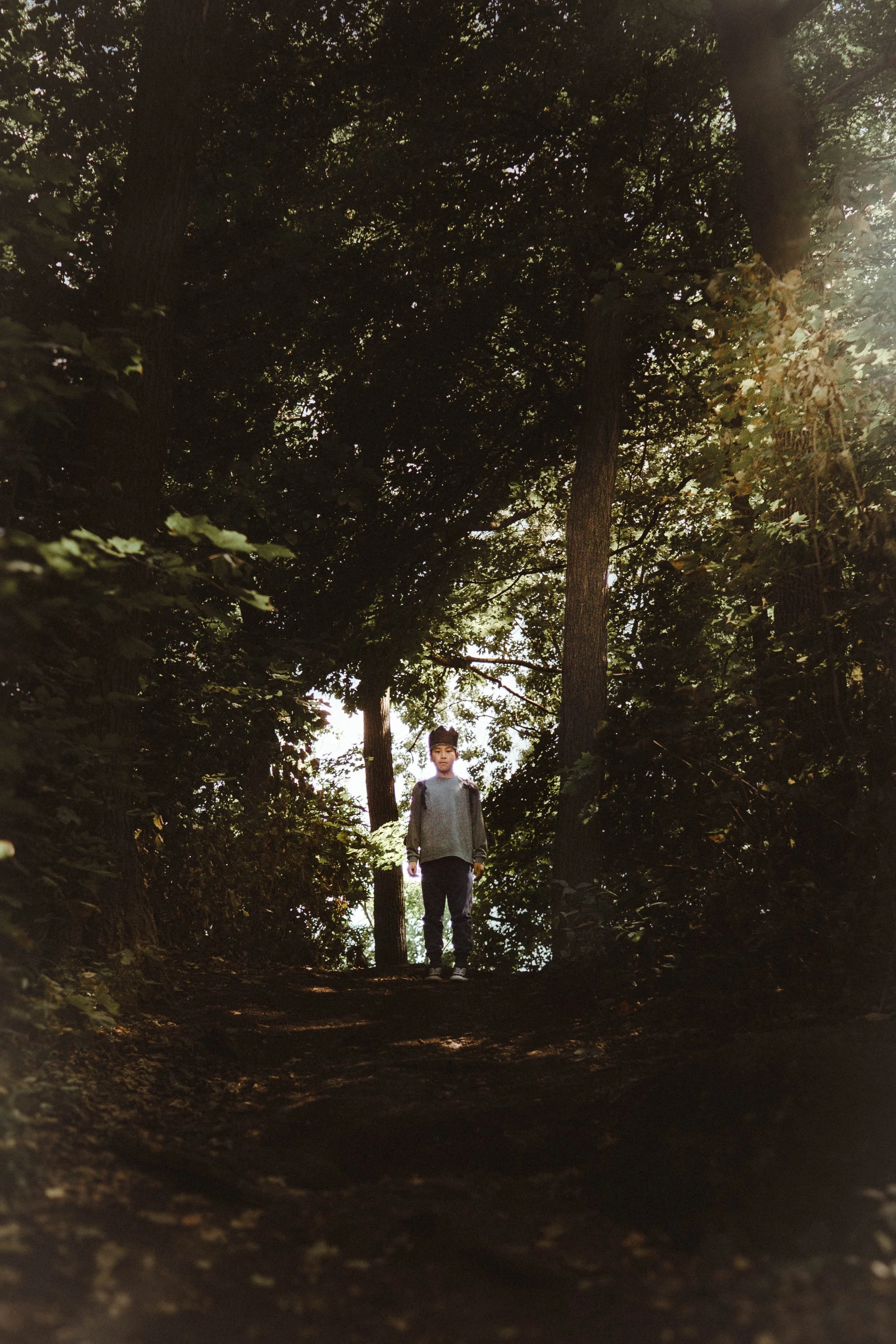 A boy standing on a wooded forest trail surrounded by tall trees, with sunlight filtering through the leaves.