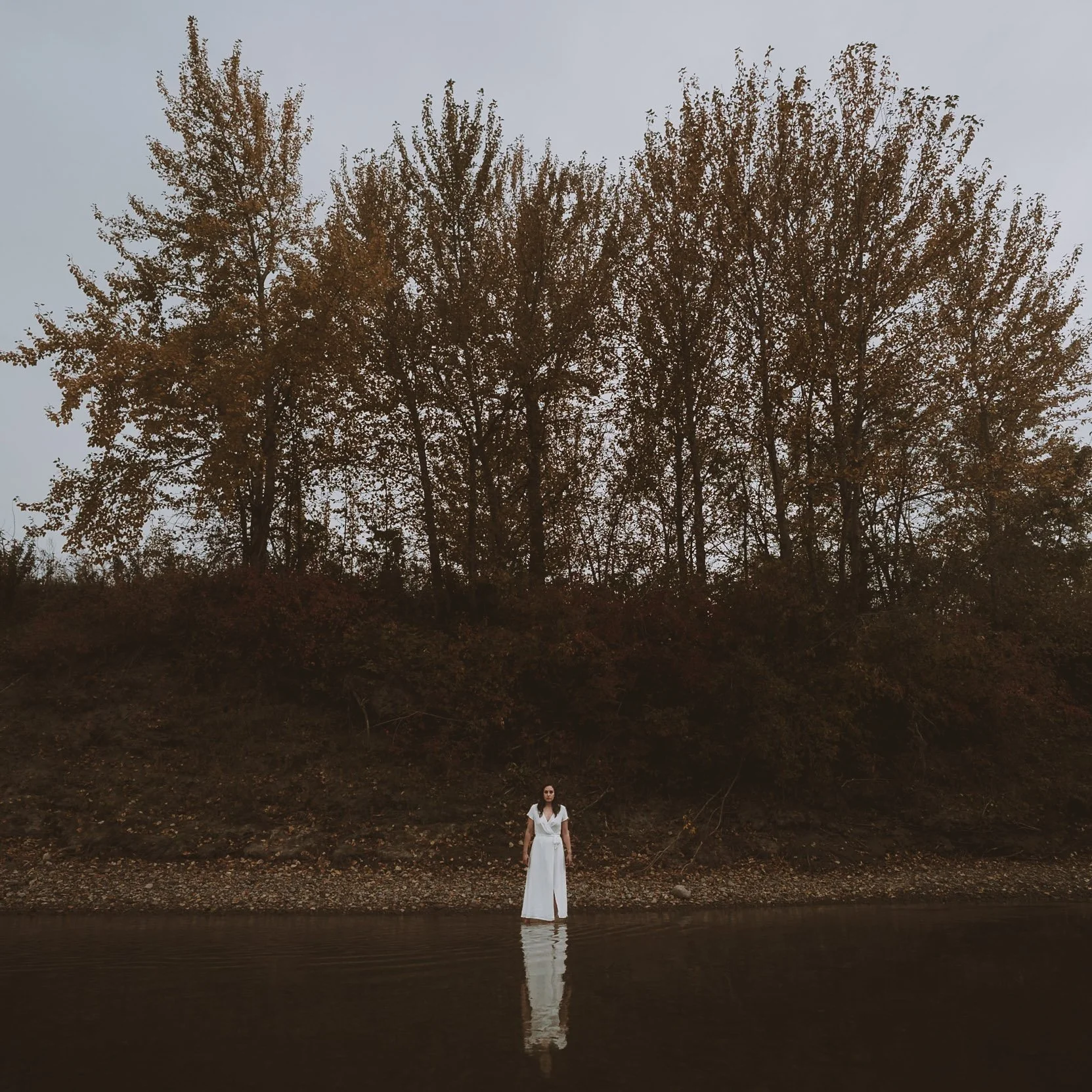 A woman in a white dress standing near a body of water with large trees in the background under a gray sky.