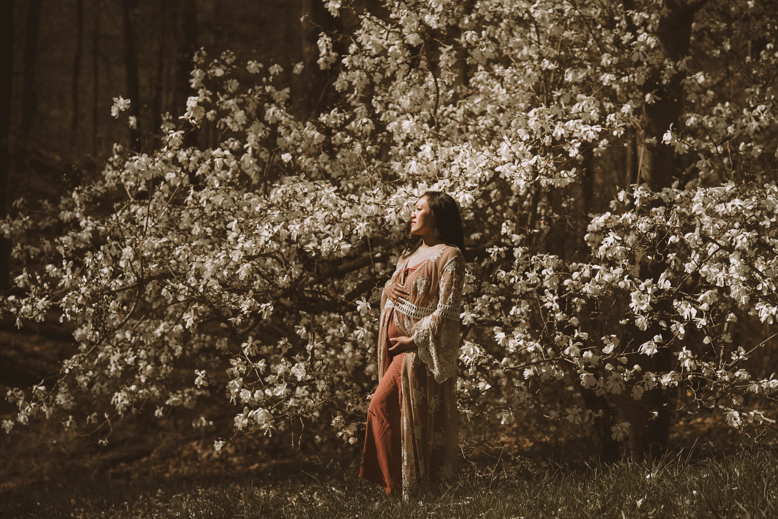 A woman standing in front of a blooming tree with white flowers, wearing a traditional dress, with her hand on her chest and eyes closed, at night.