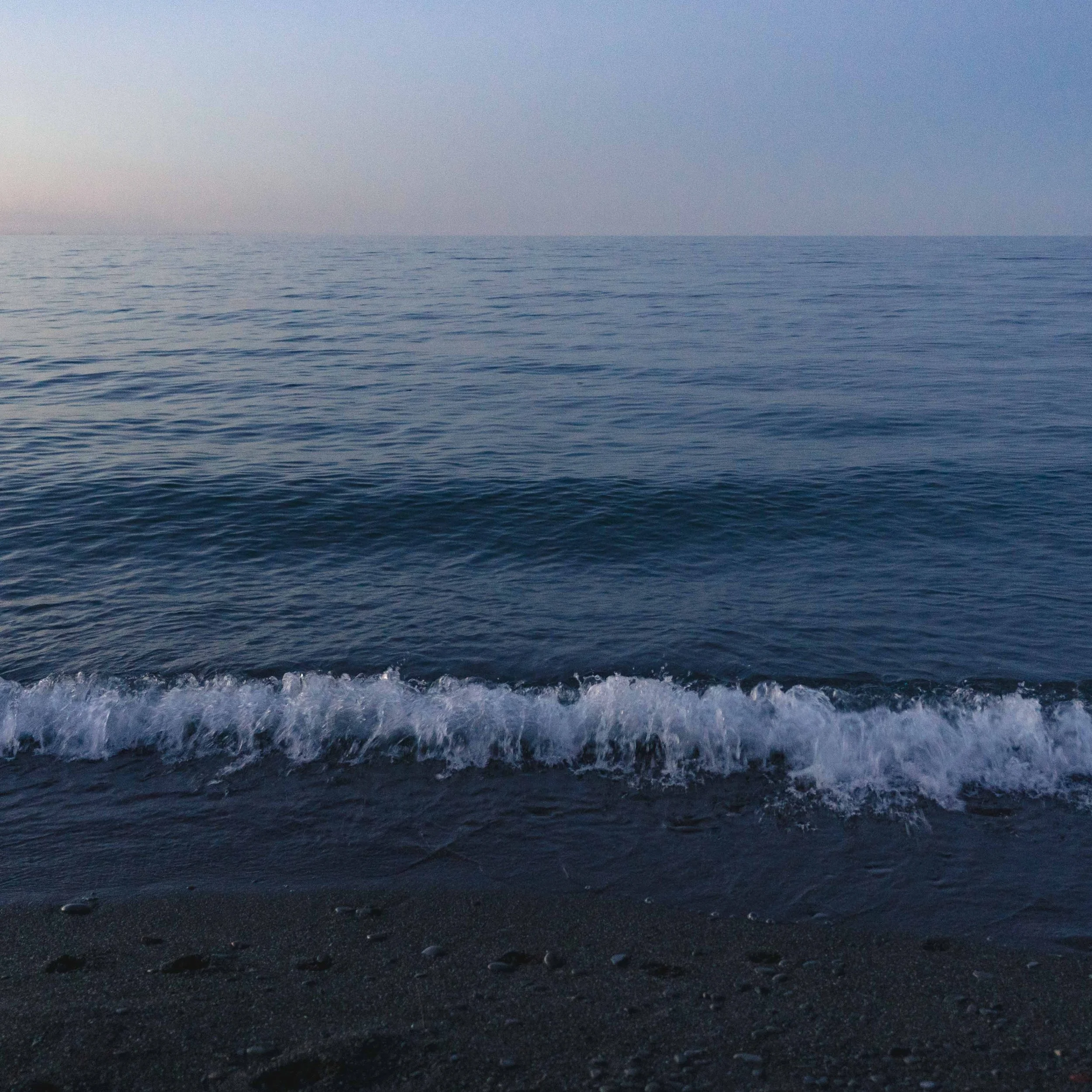 Calm ocean water with gentle waves hitting a dark sandy beach at sunset.