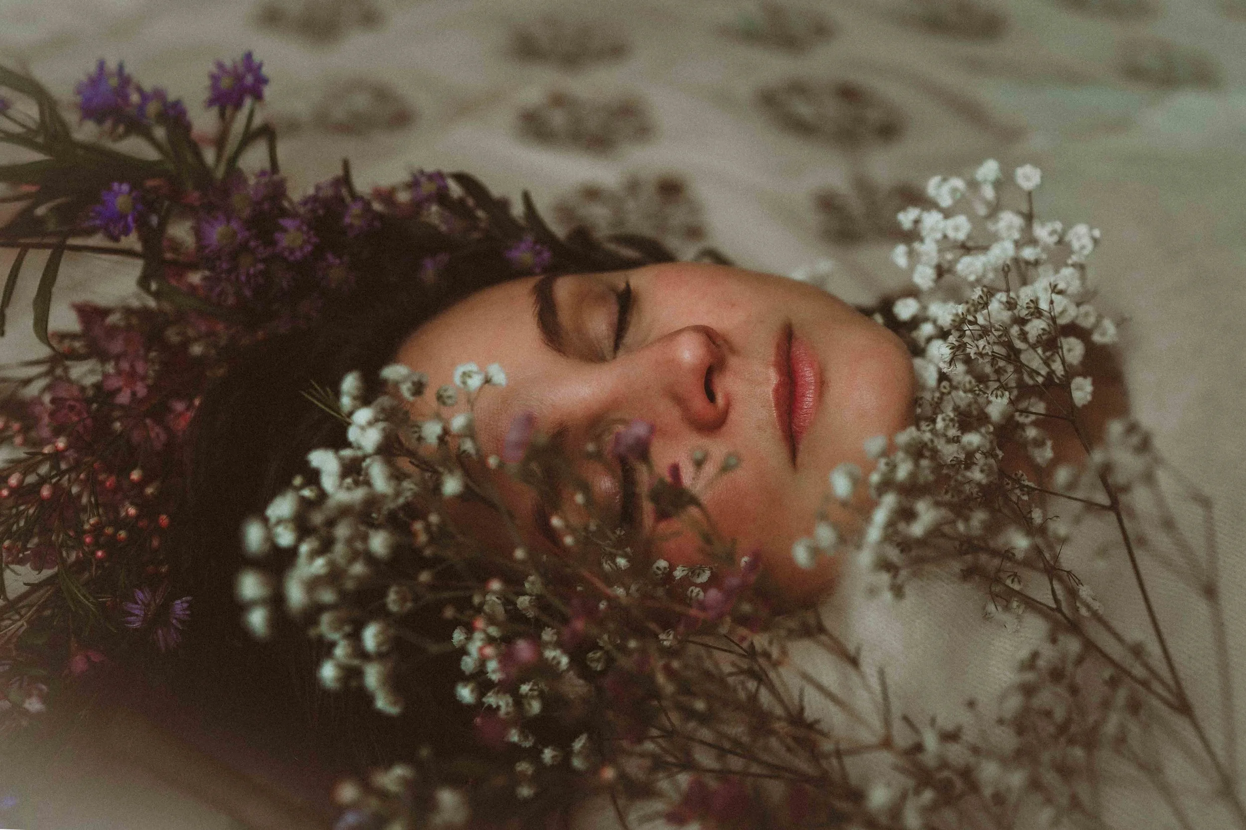 A woman with closed eyes surrounded by white, purple, and pink baby's breath flowers, lying on a light-colored surface.