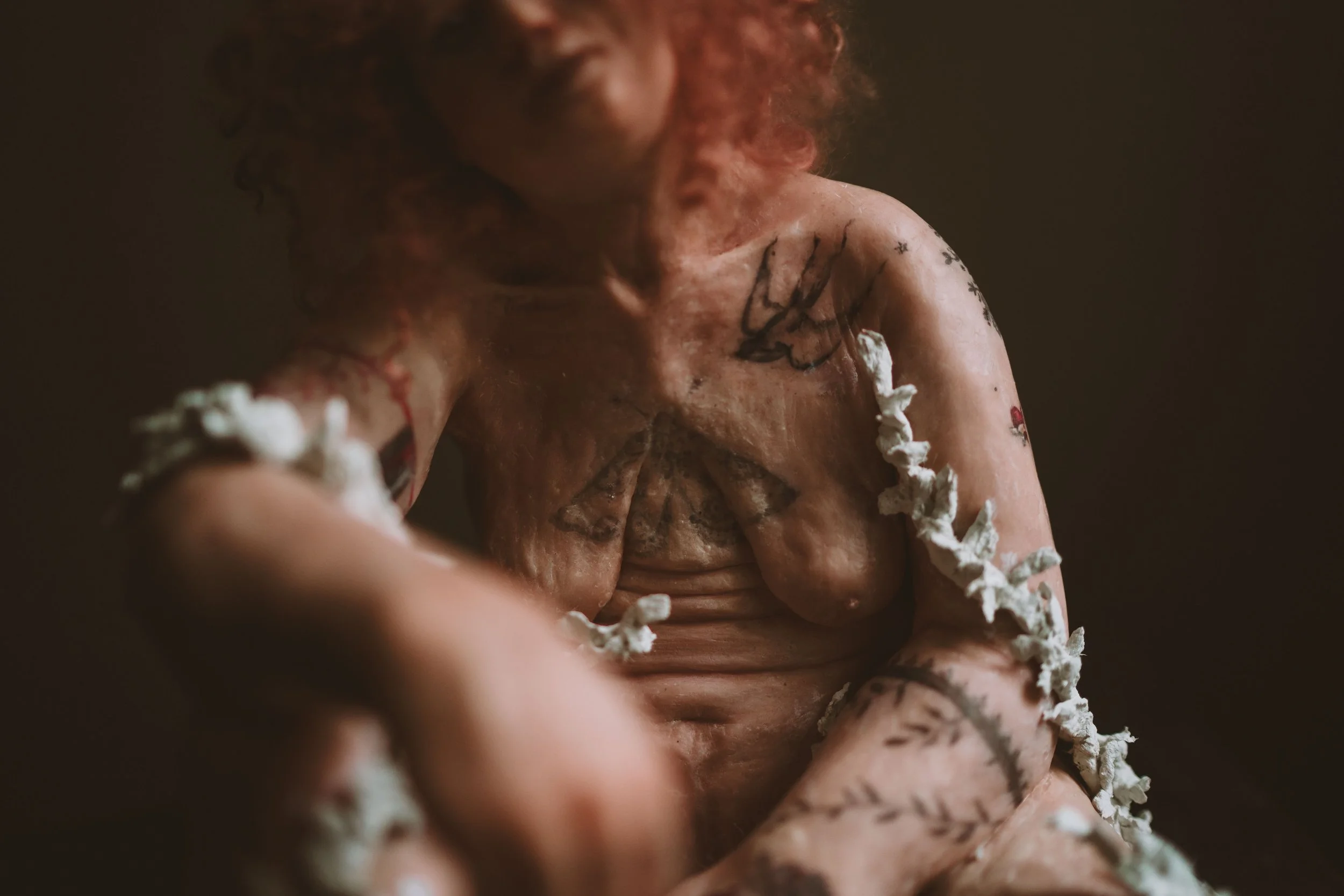 A person with curly hair covered in white foam or whipped cream, looking down, with tattoos visible on their arm and chest.