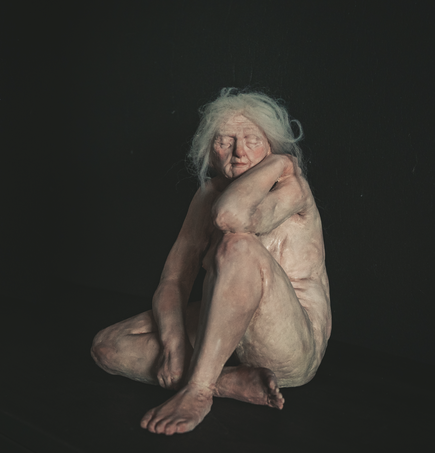 A sculpture of an elderly woman with long gray hair, sitting in a curled pose with her arms wrapped around her knees, against a dark background.