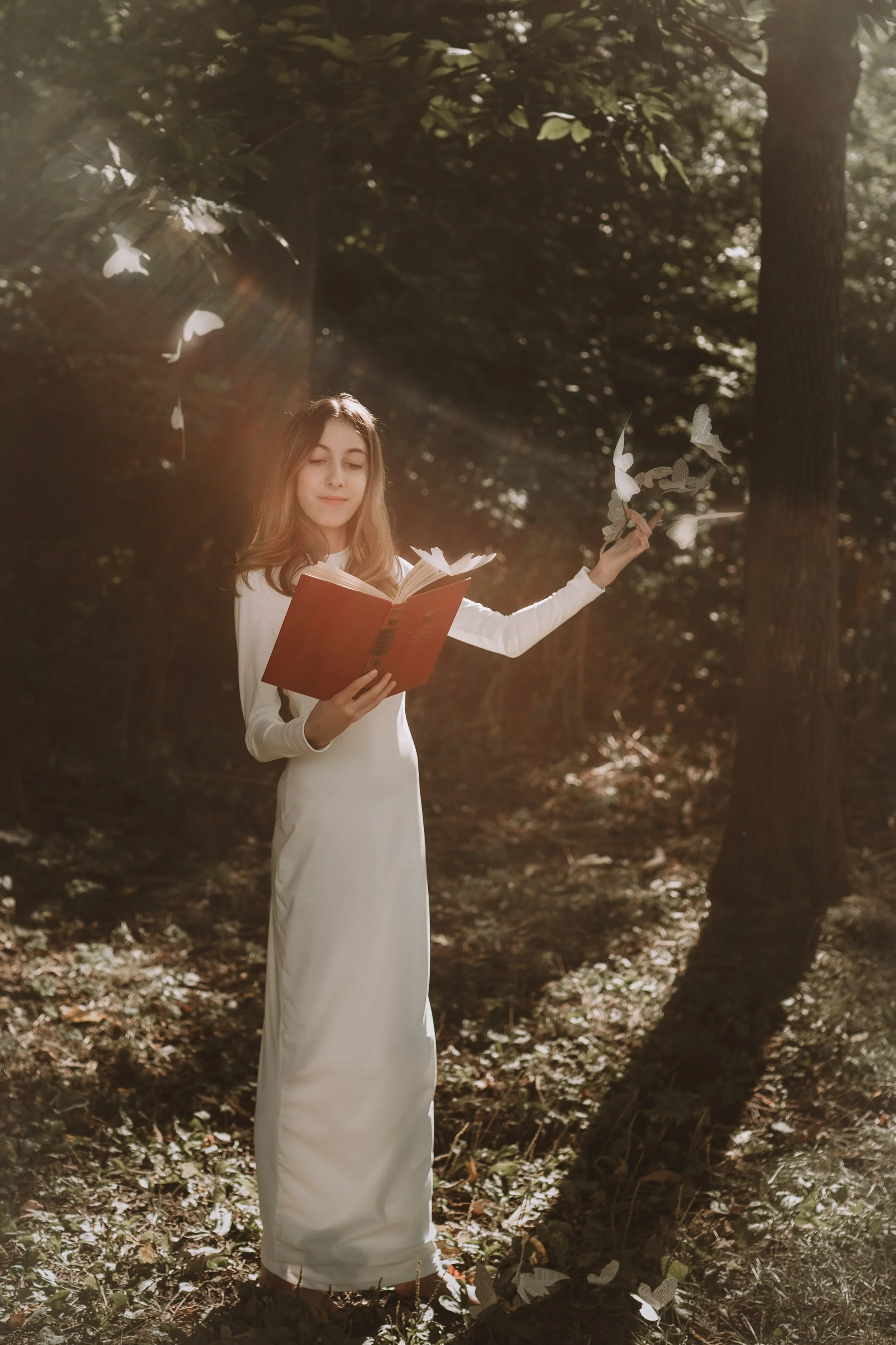 A young woman in a white dress reading a book while standing outdoors among tall trees, with sunlight filtering through the leaves.