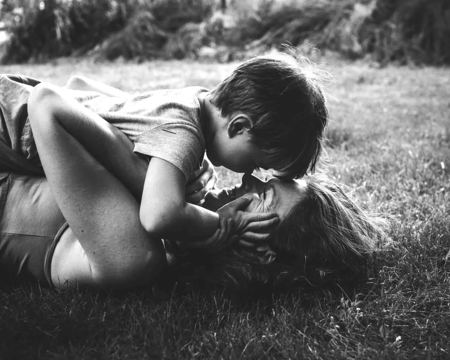 A young boy and an adult person lying on the grass, engaging in a close, affectionate moment, touching foreheads and holding faces.