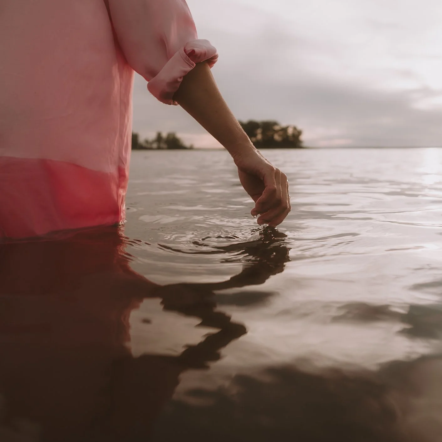 Person in pink jacket wading into water, touching the surface with their finger, during sunset or sunrise, with trees in the distance.