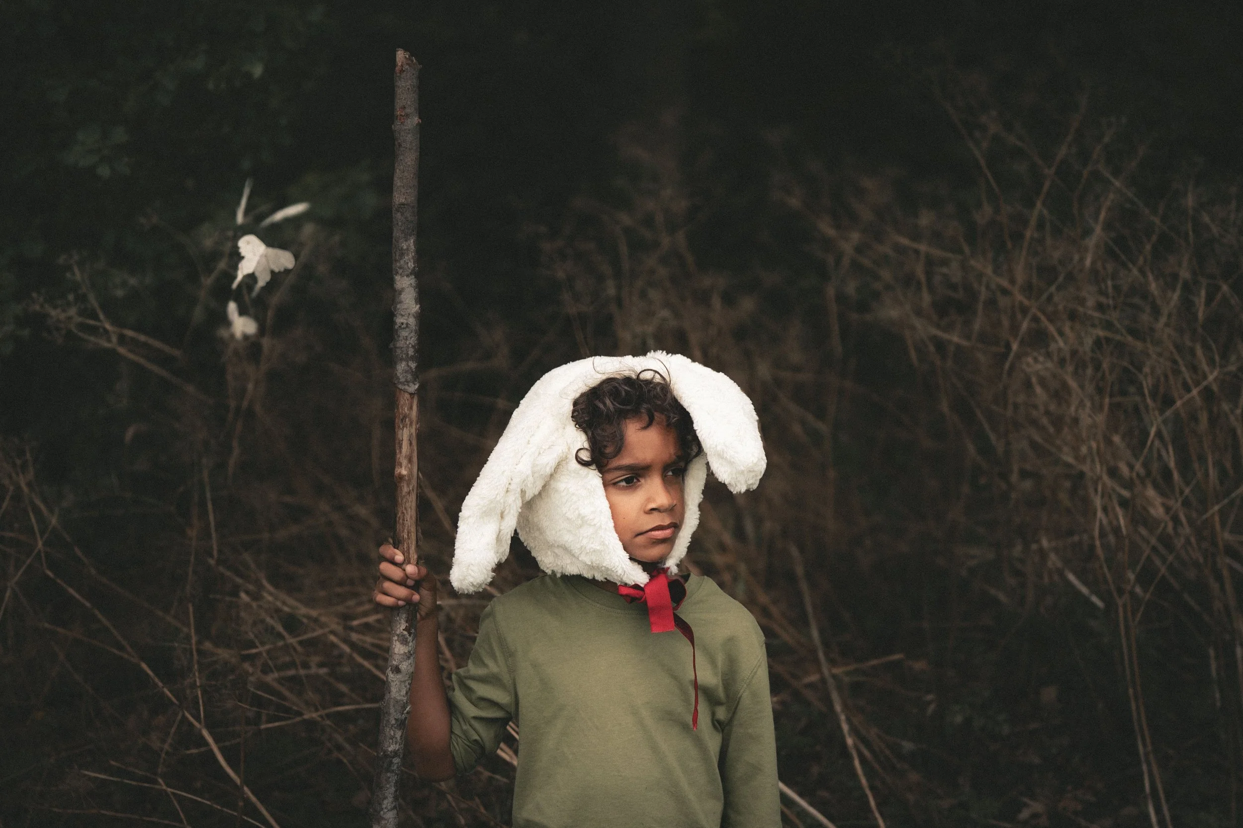 A young boy dressed in a green shirt and wearing a white fluffy bunny hat with long ears, holding a stick, standing outside in a wooded area at night.