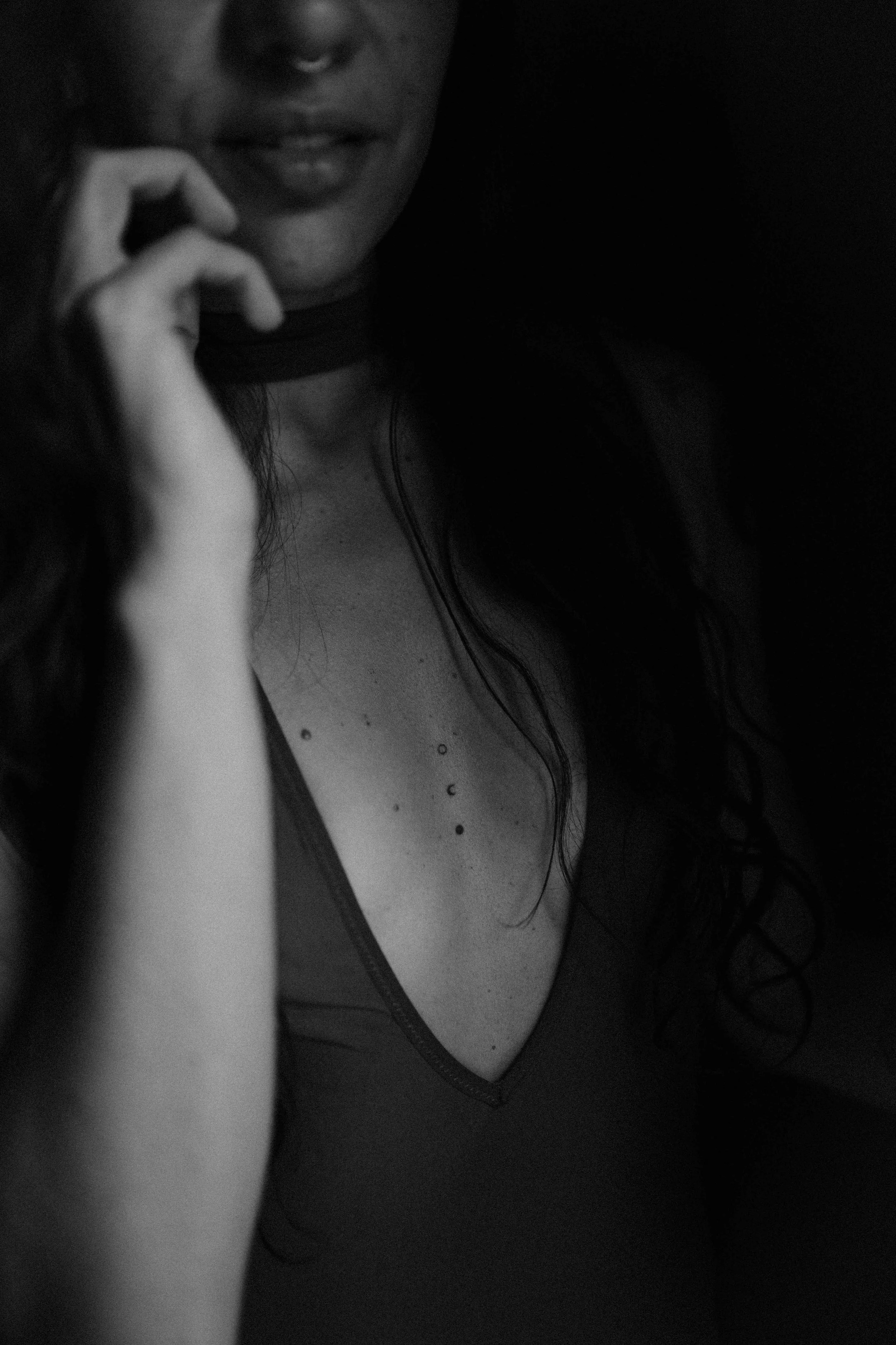 A woman with dark curly hair and multiple piercings on her chest, touching her face with her hand, partially visible smiling face, in a dark setting, black and white photo.