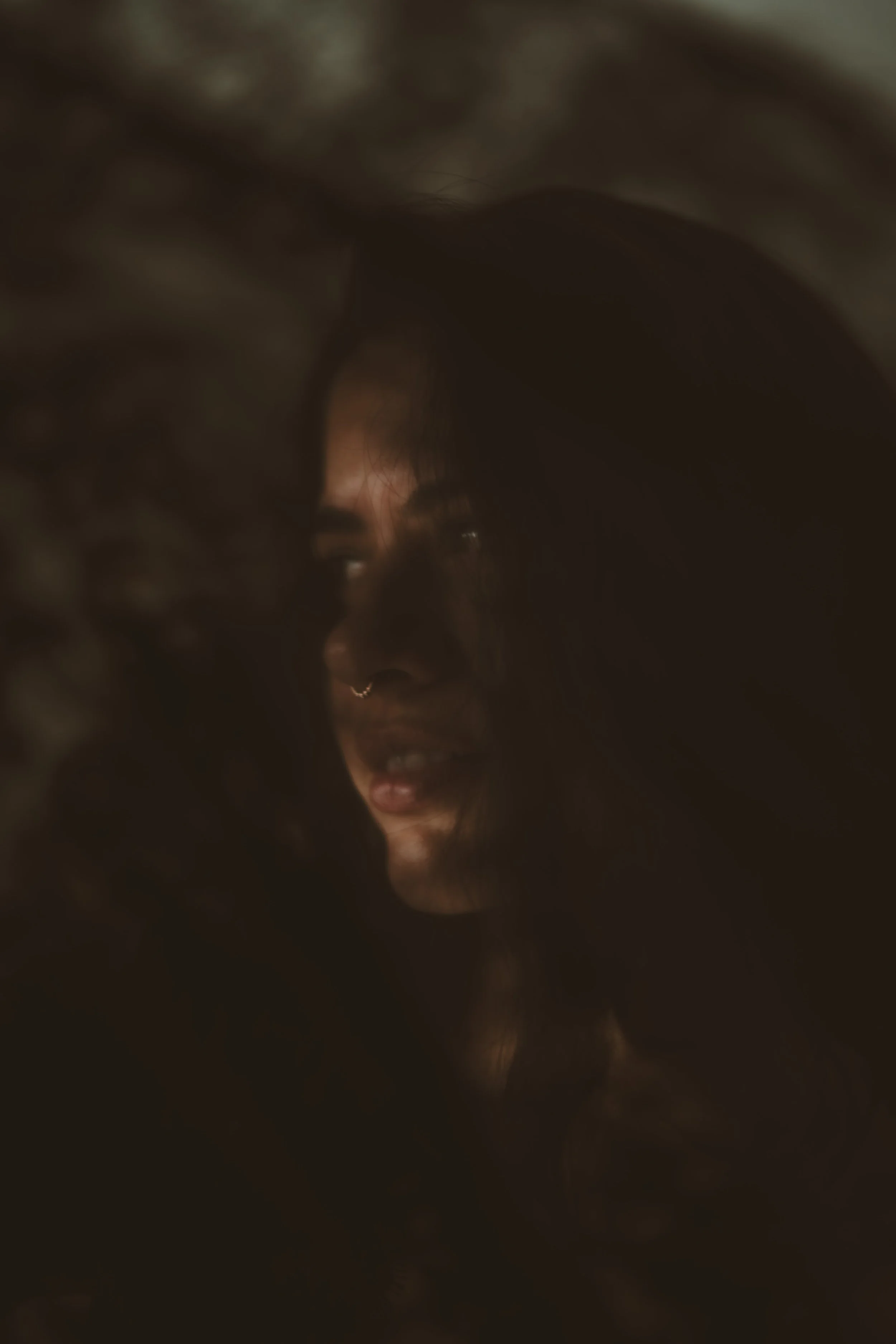 A woman with a nose ring and long hair partially obscured by a dark, textured foreground, with soft lighting highlighting her face.