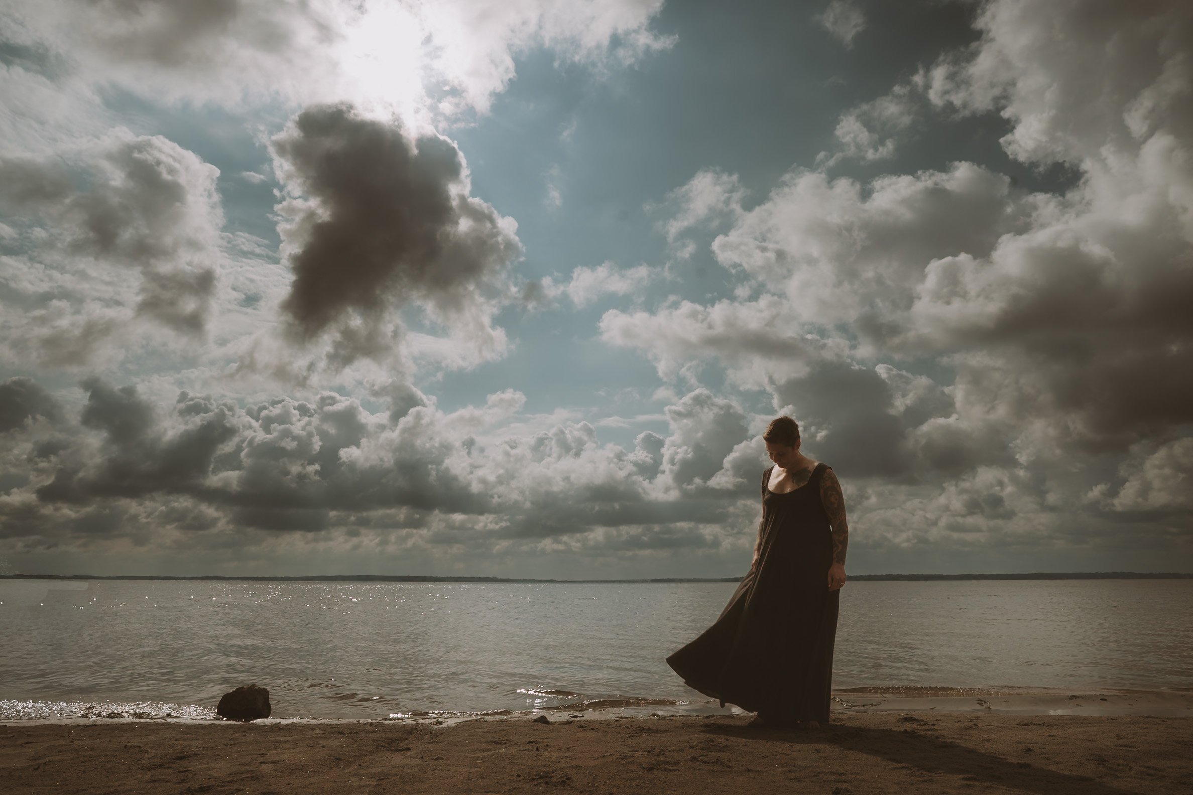 A person in a long dark dress standing on a beach near water, with cloudy sky overhead.