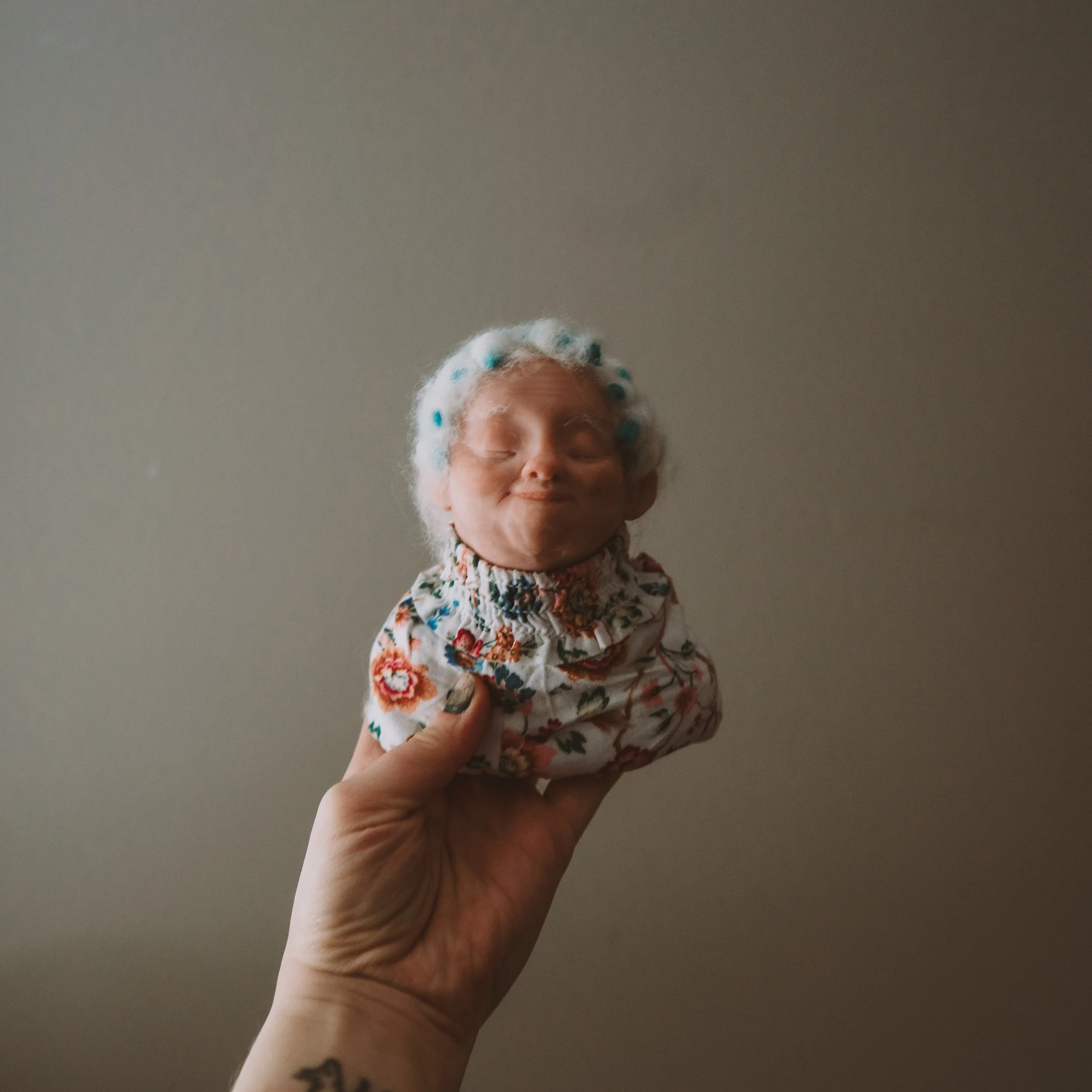 A person holding up a doll with closed eyes and a white bonnet with blue polka dots in front of a gray wall.