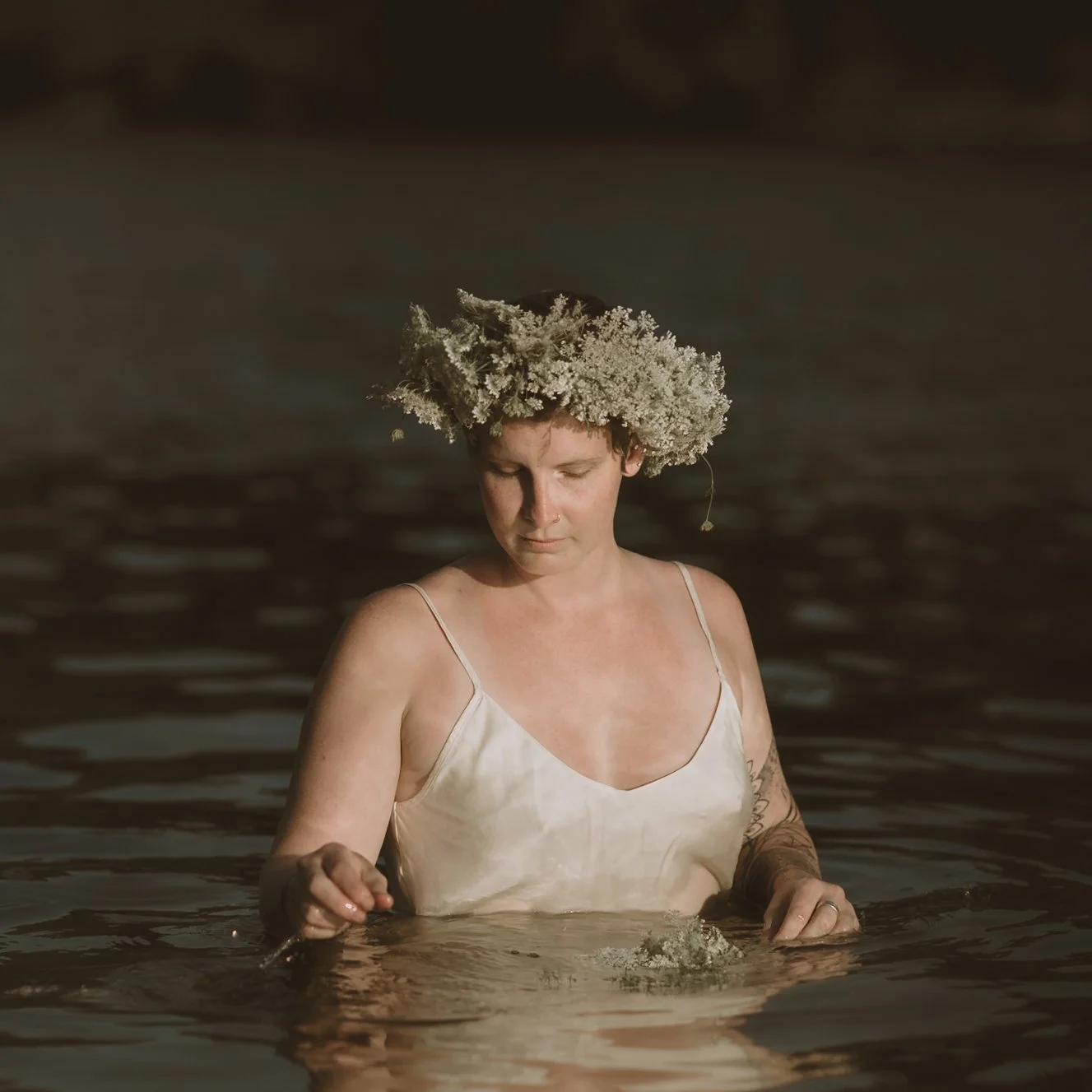 Woman standing in water with a floral crown, wearing a satin dress, looking down