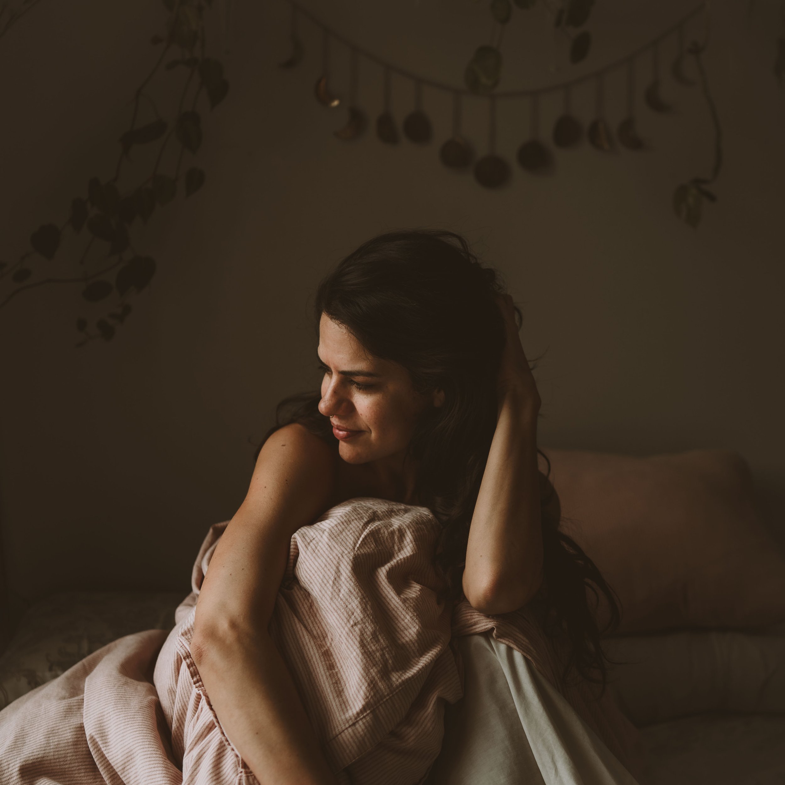 A woman sitting on a bed, smiling softly with her eyes closed, resting her head on her hand, in a dimly lit room with floral wall decorations.