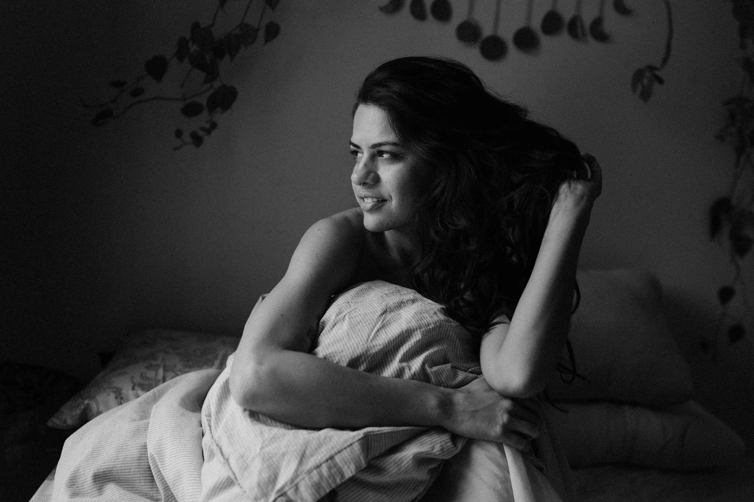 A young woman with dark, wavy hair and a nose ring resting on a bed, looking off to the side with a slight smile, in black and white.