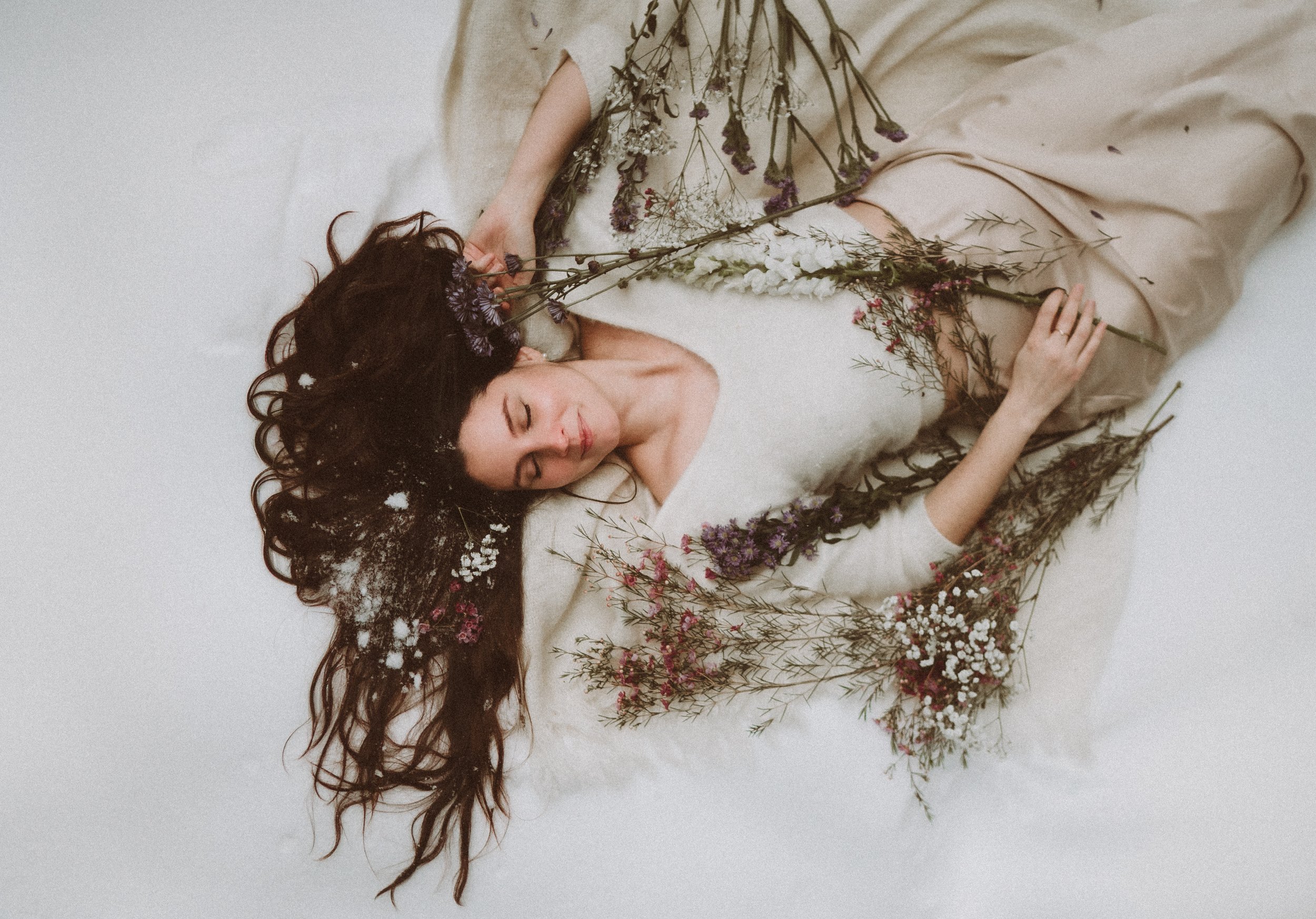 A woman with long, dark, curly hair lies on a white surface, eyes closed, holding flowers near her face, surrounded by various dried flowers and plants.