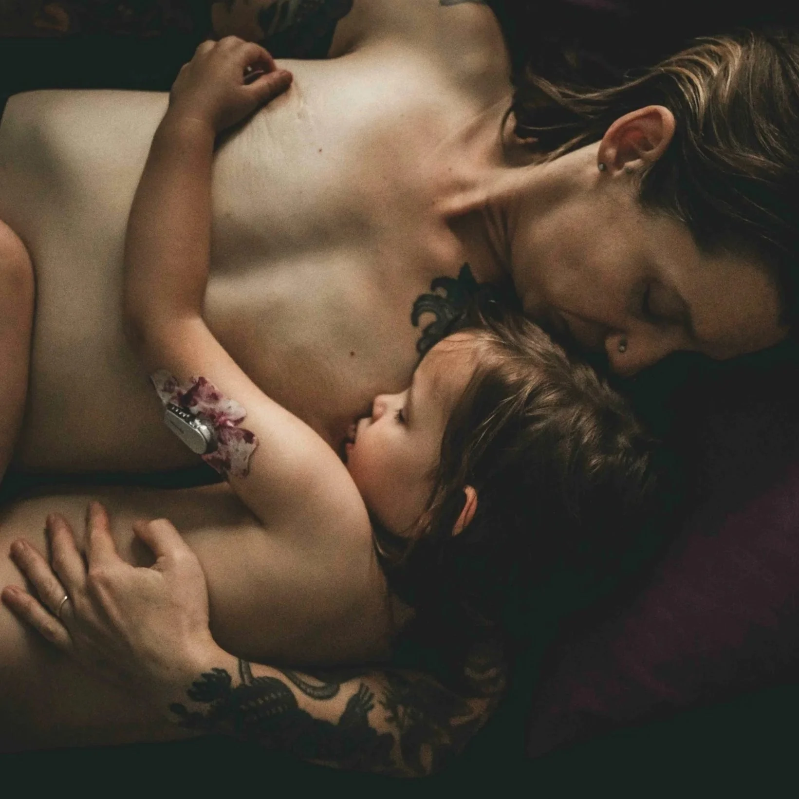 A woman with long dark hair and earrings lying shirtless on her side with a young girl leaning on her, hugging her, both smiling. The girl's arm has a temporary tattoo and a wristwatch.