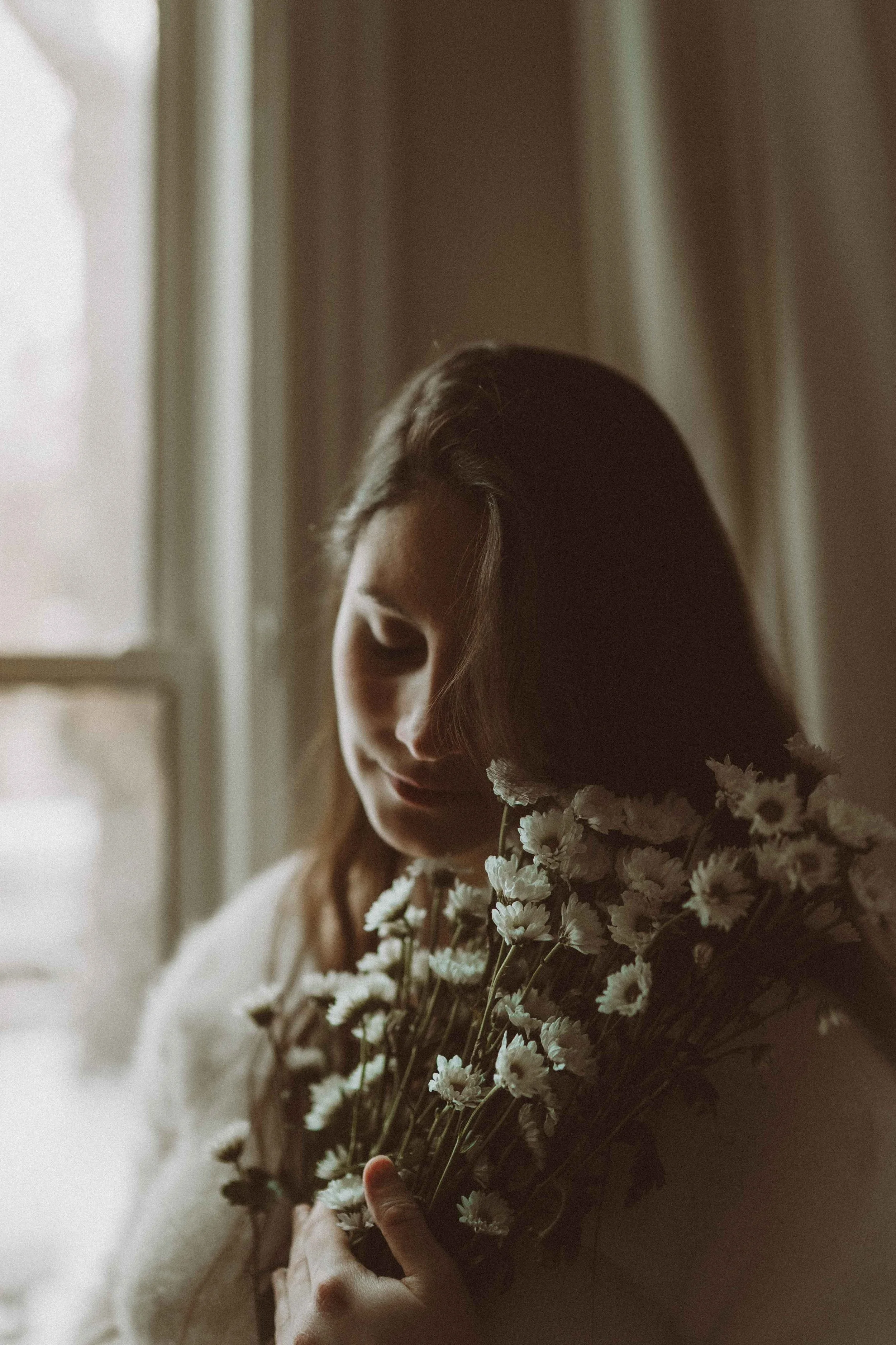 A woman holding a bouquet of white daisies near her chest indoors, near a window with curtains, with a soft, warm light illuminating her face.