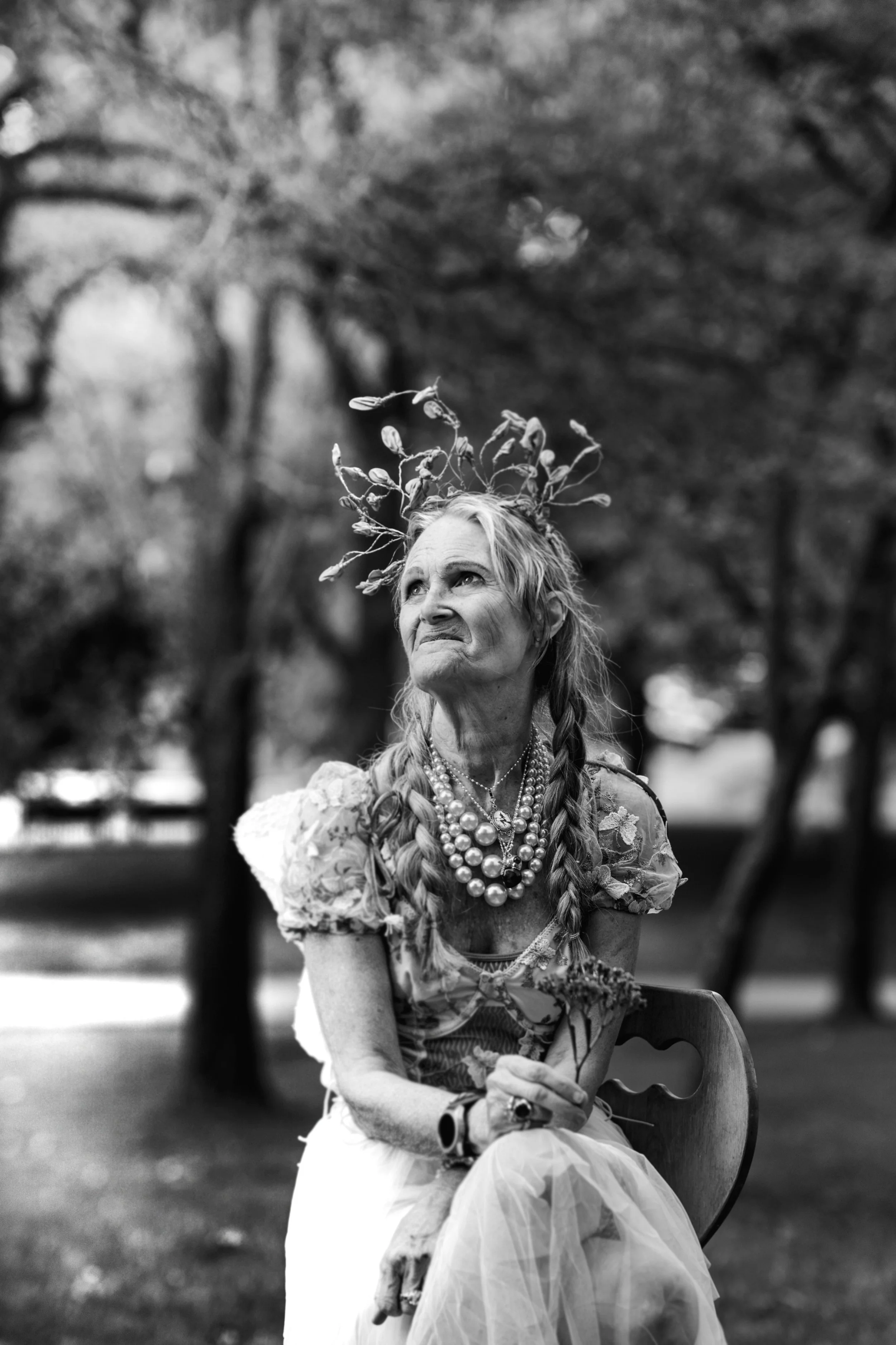 A woman with long braids and multiple pearl necklaces, sitting outdoors on a wooden chair, wearing a floral top and tulle skirt, with a crown made of twigs or branches on her head. The background shows trees and a natural setting, and the photo is in