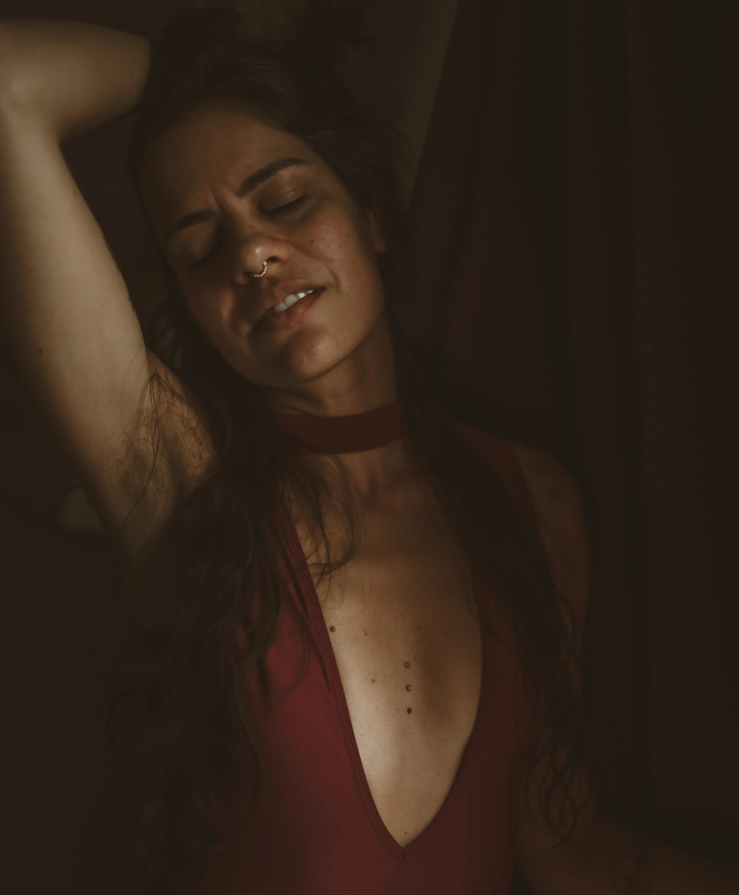 A woman with long dark hair, freckles, and multiple facial piercings, wearing a deep red top with a choker neckline, appears to be in a moment of emotional expression with her eyes closed and her head tilted back.