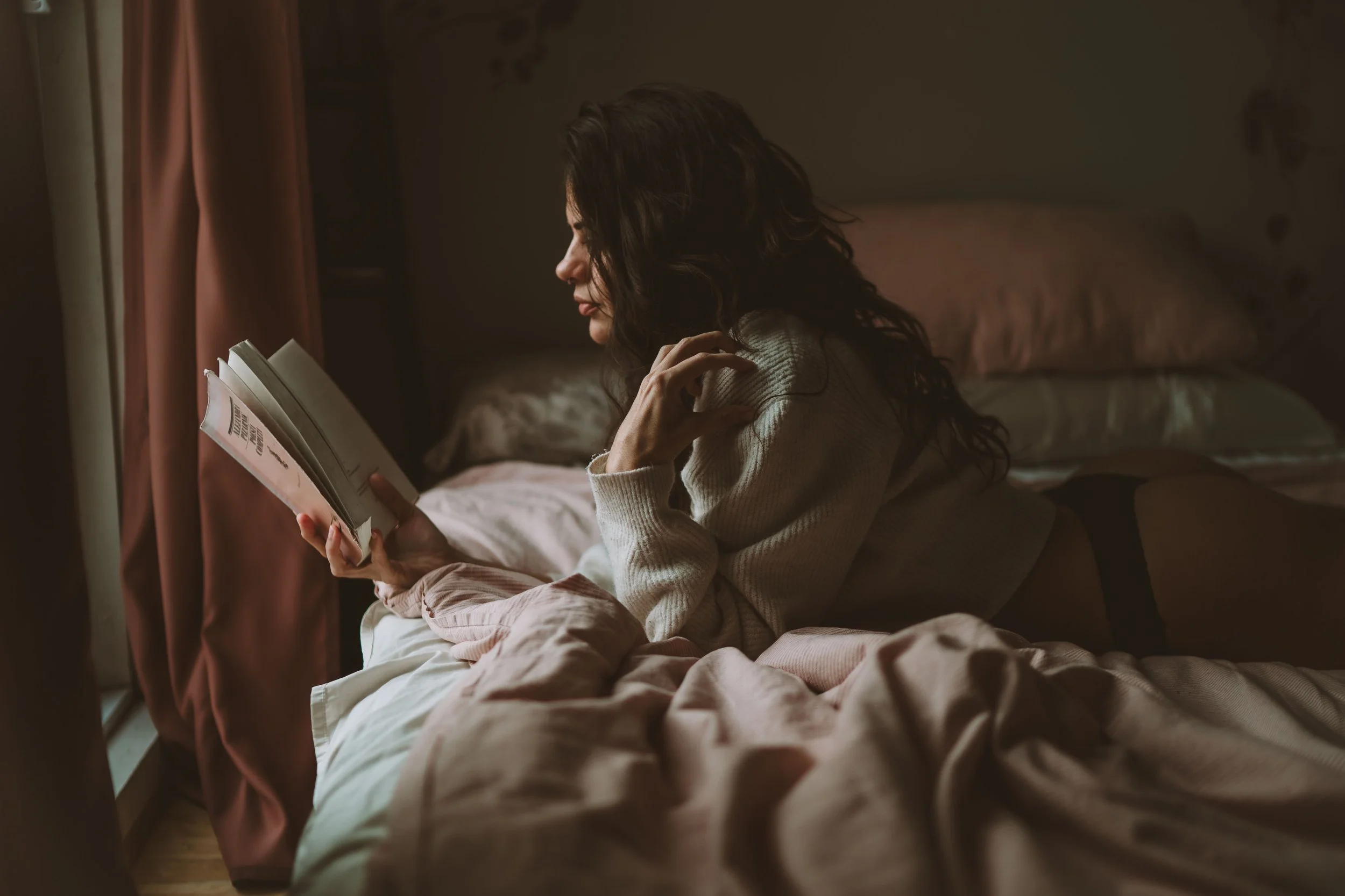 Woman with long dark hair lying on bed, reading a book near window with pink curtains, in a dimly lit room.