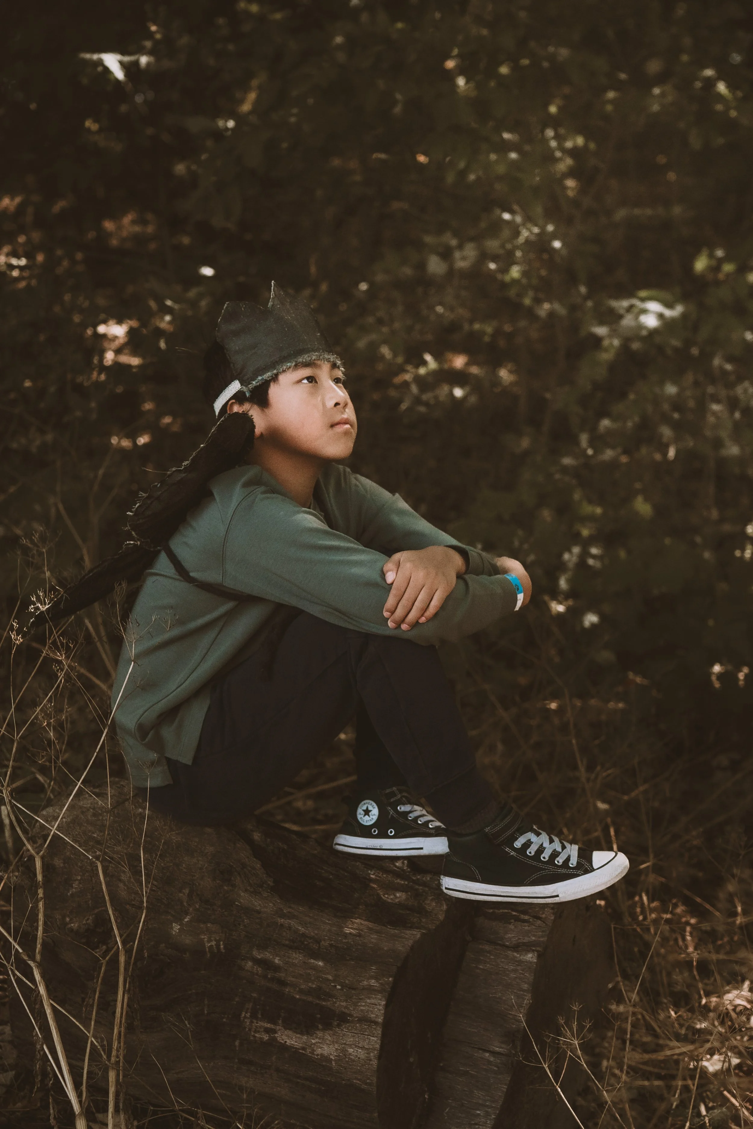 A young boy in a gray jacket, black Converse sneakers, and a gray hat sits on a large tree branch in a wooded area at dusk, looking contemplative.
