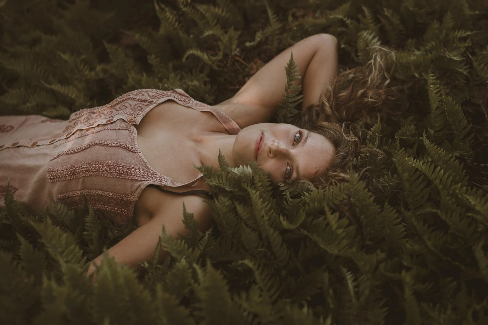 A woman with curly hair lying on her back among green ferns, wearing a pink embroidered dress and smiling slightly.