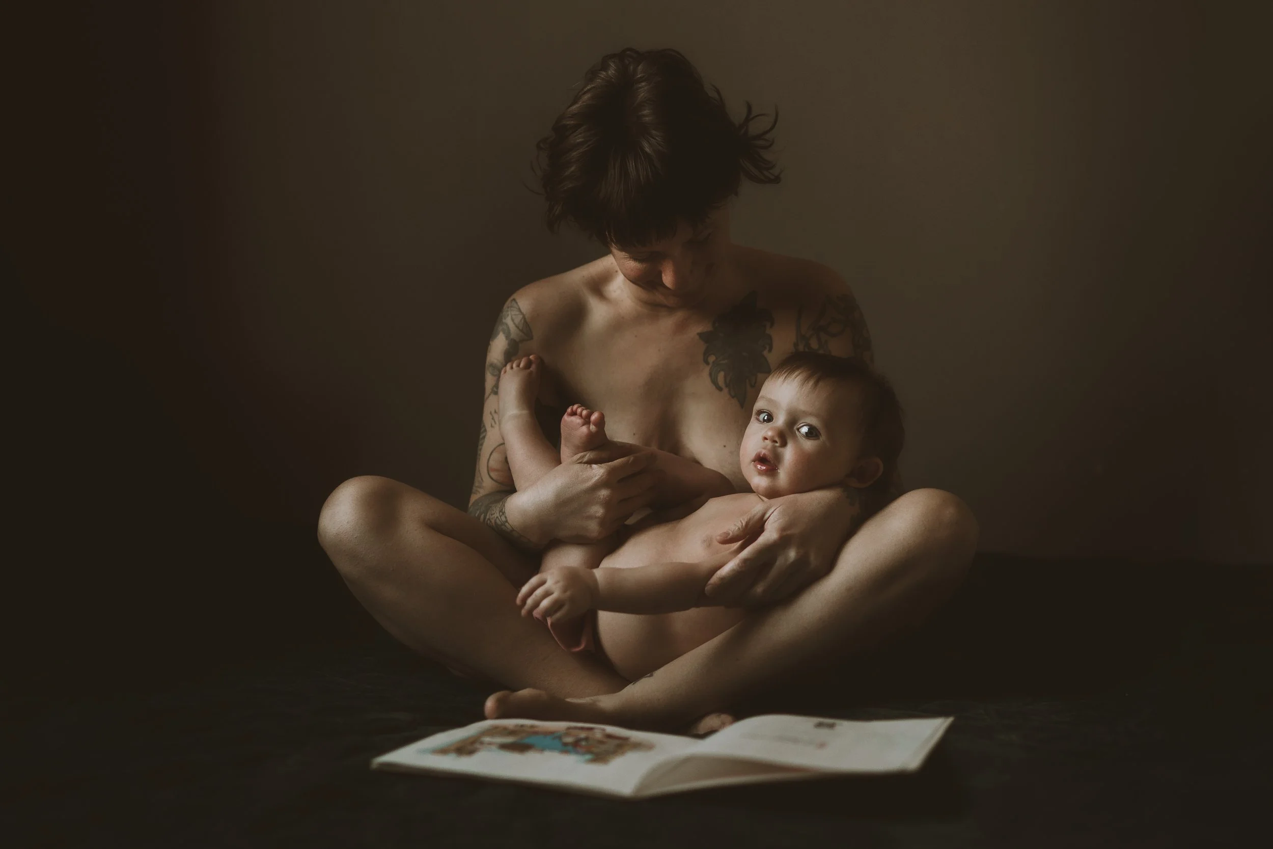 A person with short dark hair, tattoos on their shoulders and arms, sitting cross-legged in a dimly lit room, holding a baby with curly hair and looking at a magazine or book on the floor.