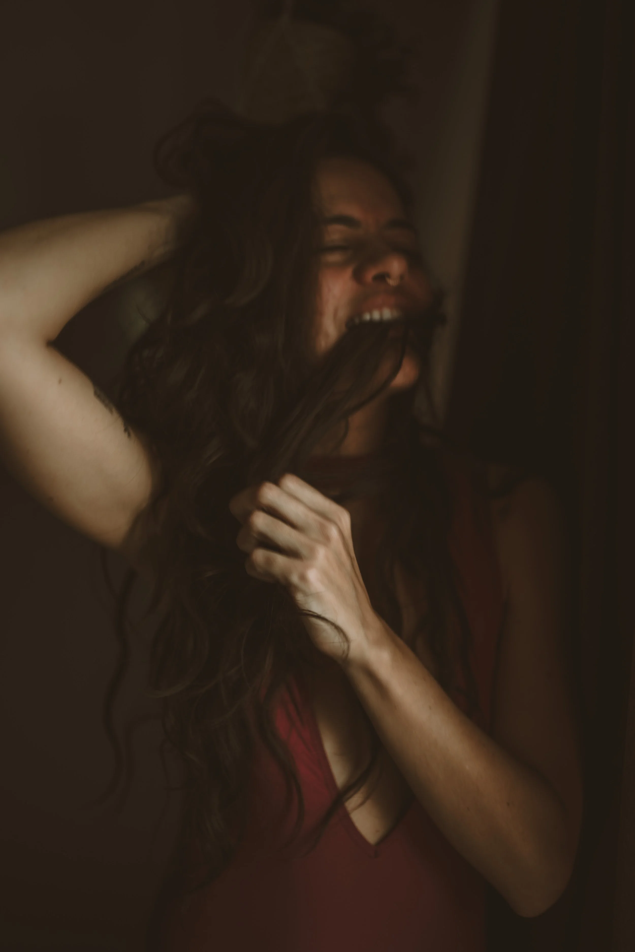 A woman with long dark hair, closed eyes, and a nose ring, holding her hair with one hand while crying or expressing intense emotion, wearing a red top, in a dimly lit environment.