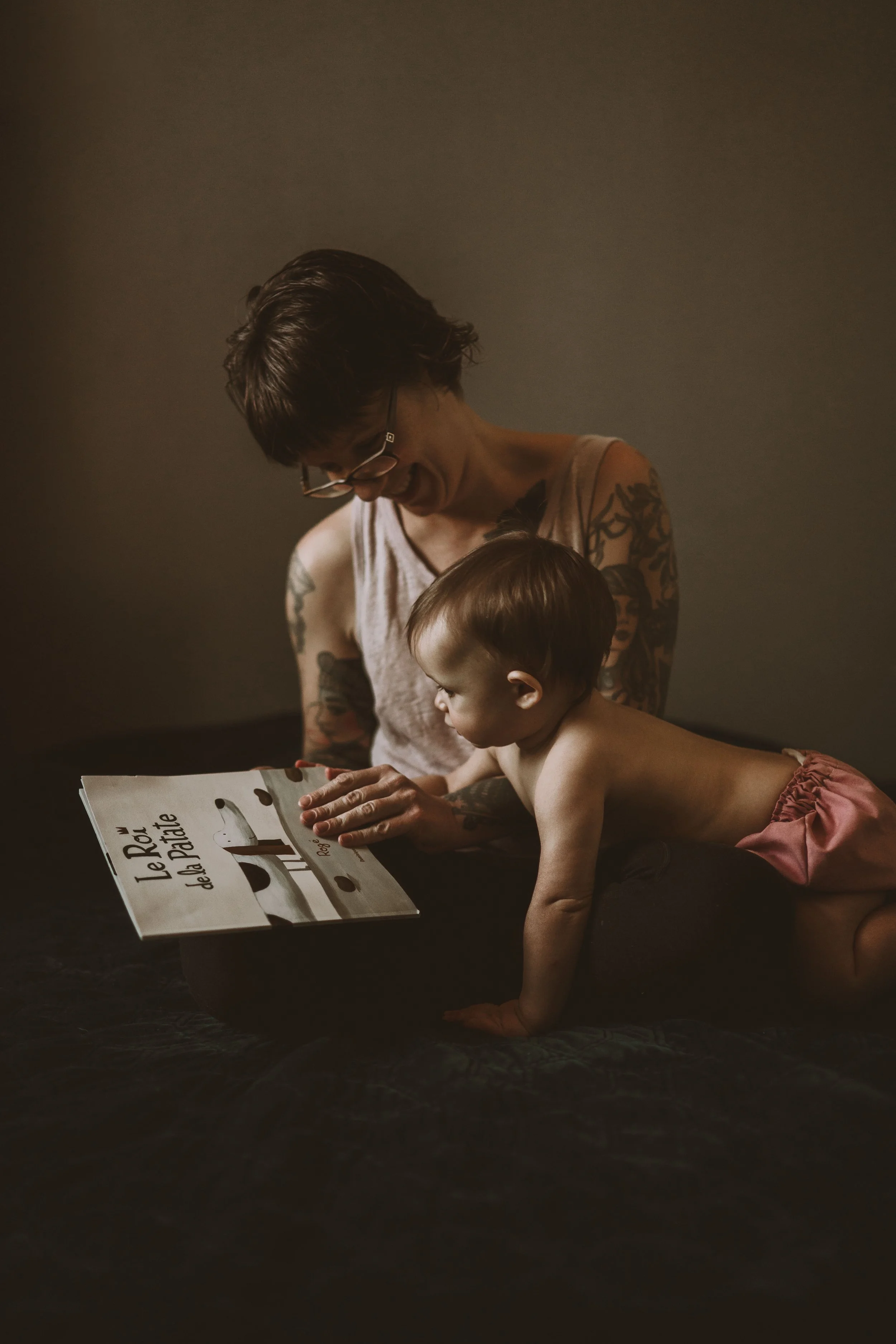 A woman with short dark hair and glasses, with tattoos on her arms, is sitting and reading a book titled 'Le Révél del de la Patate' to a young girl in pink shorts who is lying on her stomach on the floor.