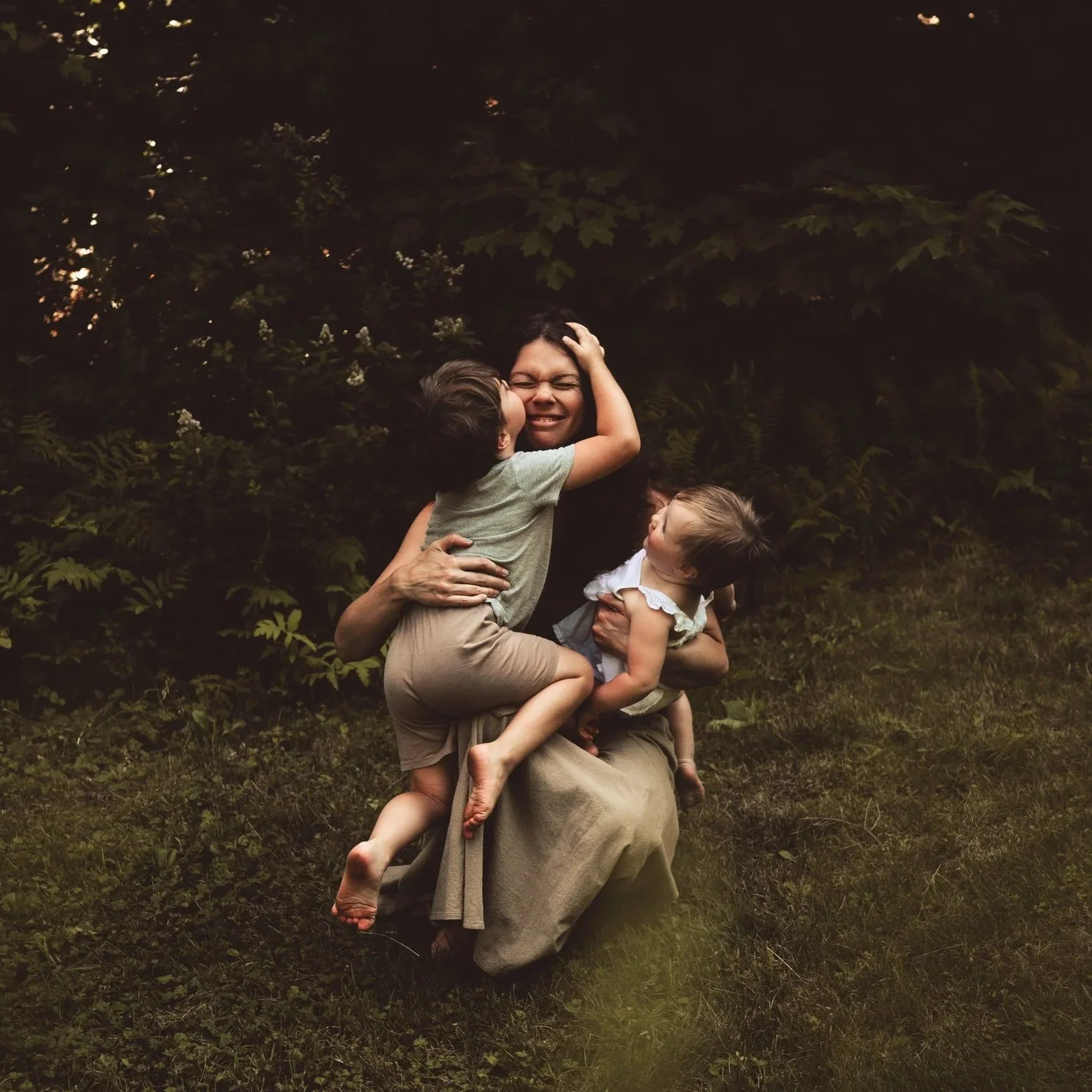 A woman with dark hair holding two young children in a grassy outdoor setting at dusk, surrounded by trees and greenery.