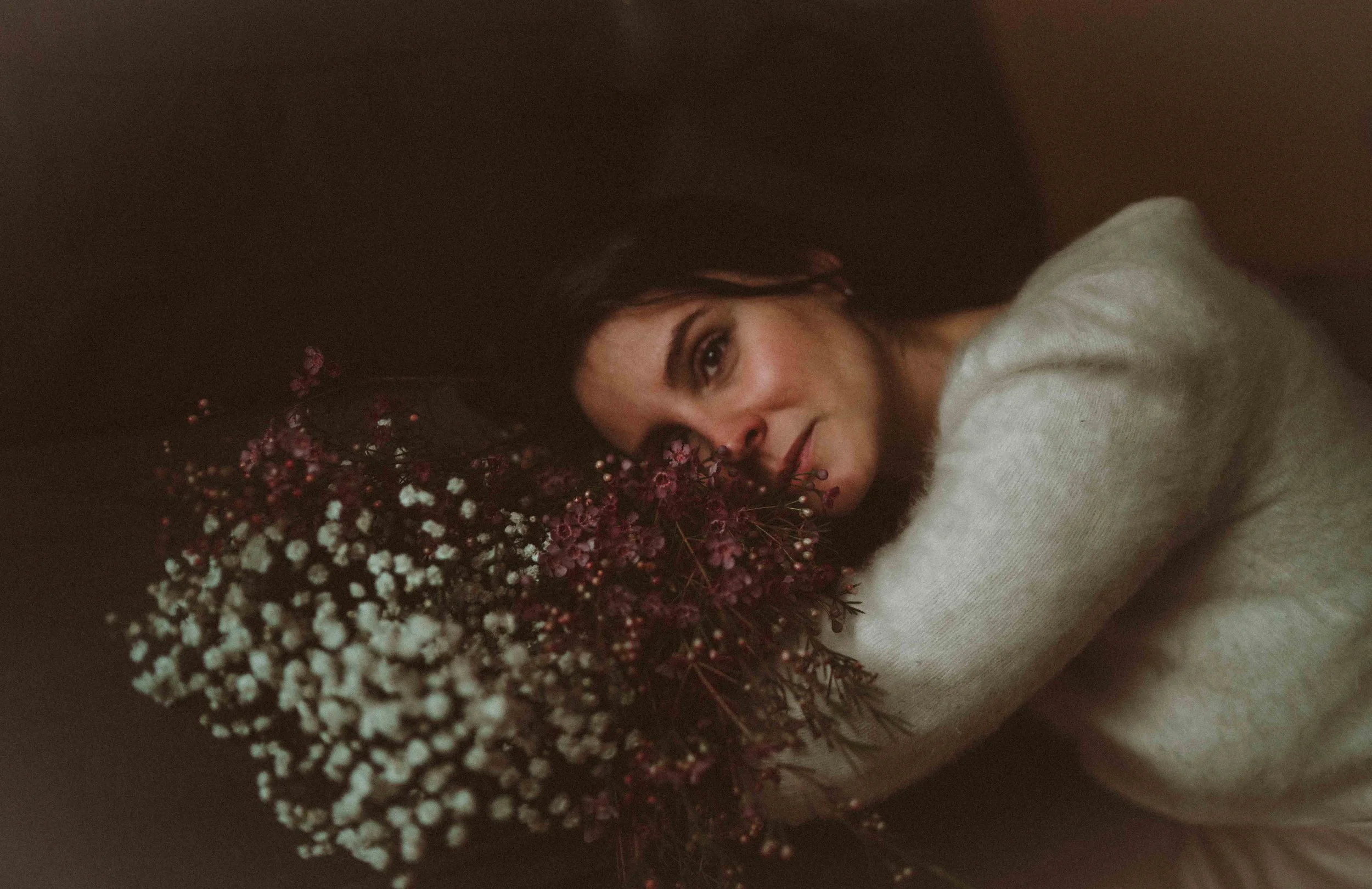 A woman with dark hair and a white sweater lying down on her side, resting her head near a bouquet of pink and white flowers, looking at the camera.