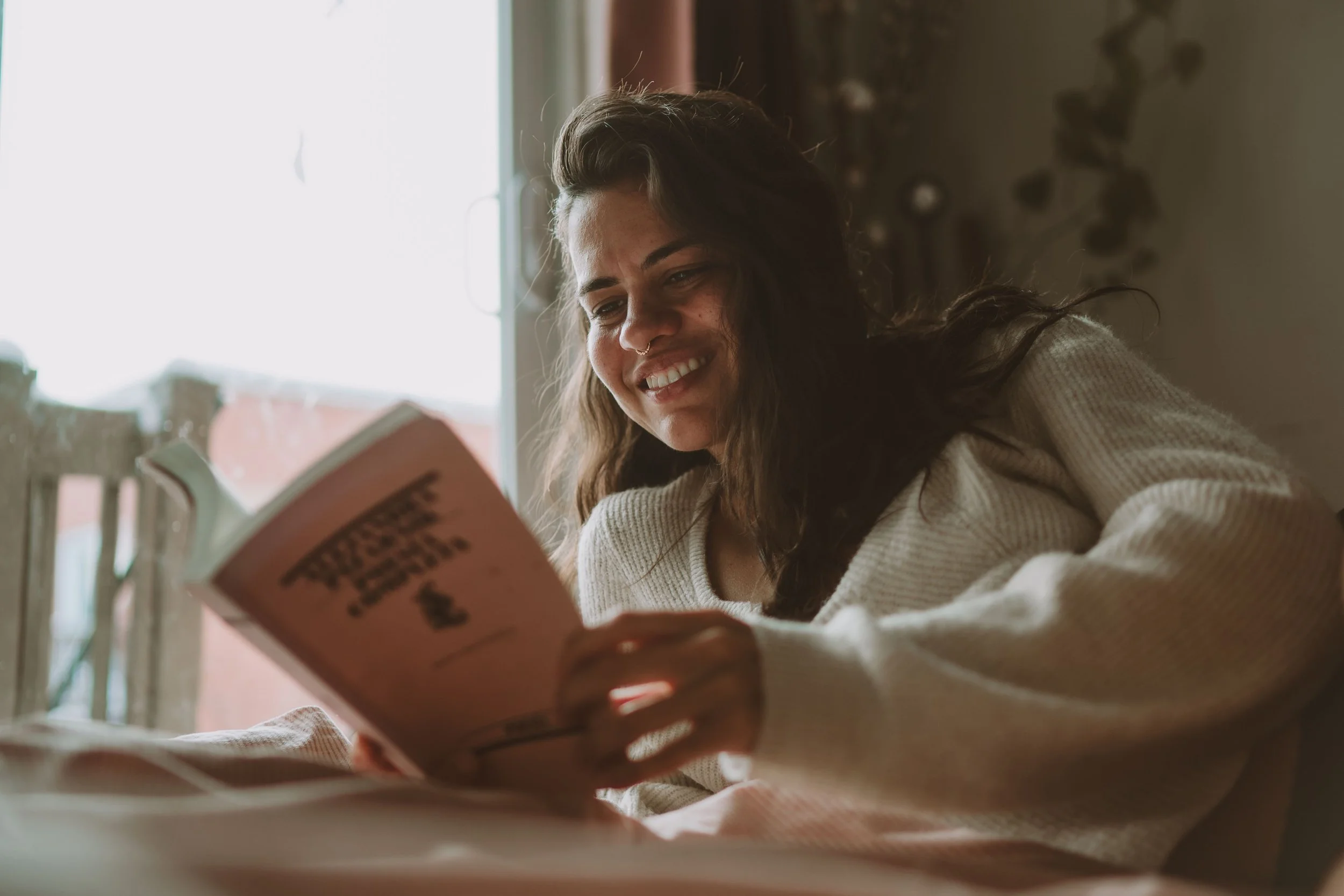 A woman with wavy dark hair and a nose piercing, smiling as she reads a book, sitting near a window with sunlight streaming in.