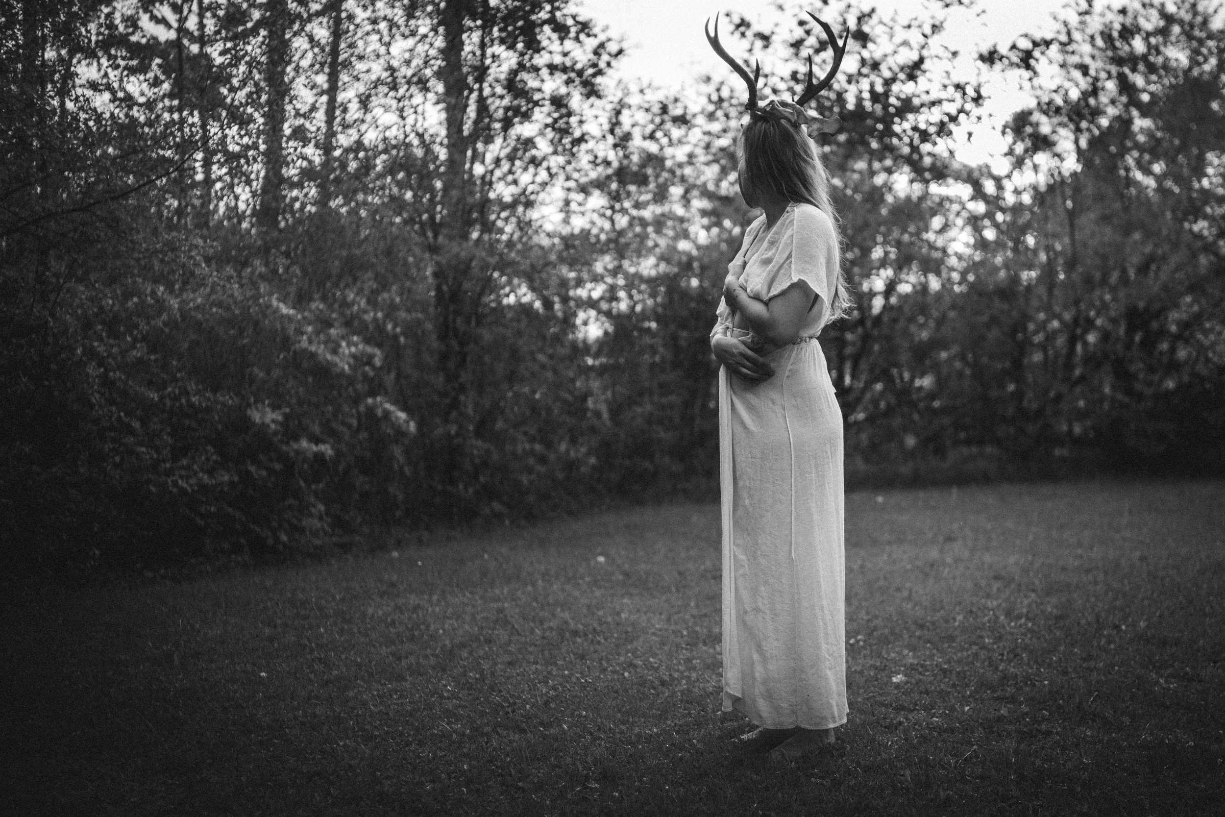 A person wearing a white dress with a deer skull and antlers on their head, standing in a forested area with trees and grass, captured in black and white.