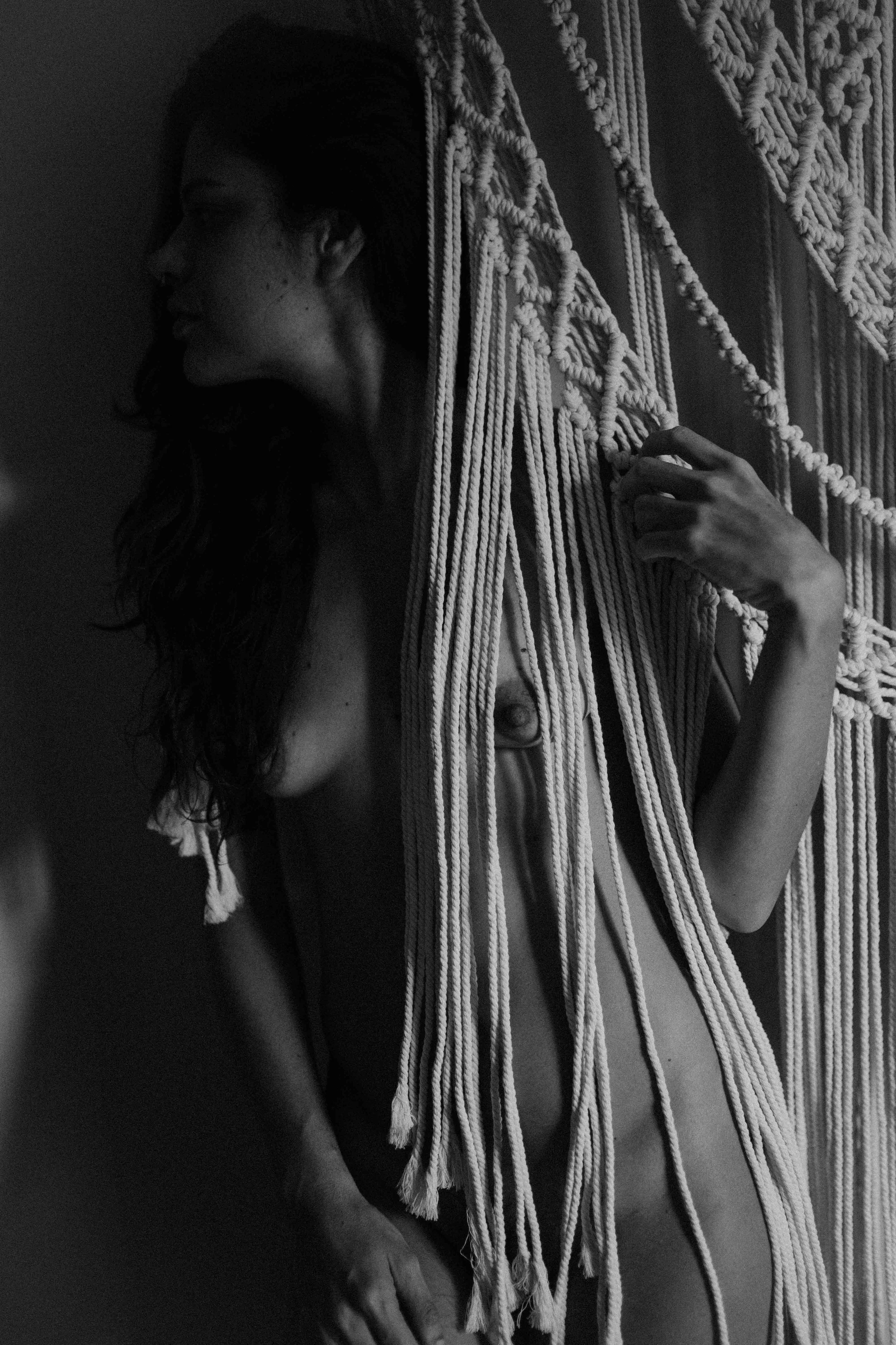 Black and white photo of a woman with long hair, partially nude, holding or touching macramé wall hanging with her right hand, with her face turned to the left side.