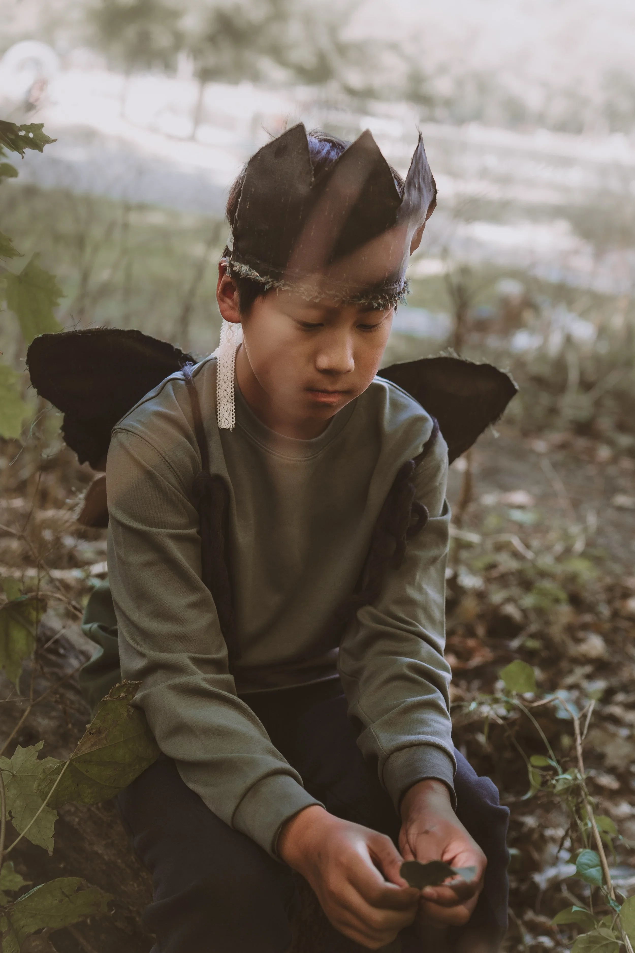 A young boy in a forest setting is sitting on a fallen log, looking down. He is wearing a brown paper crown, a gray shirt, and dark pants. His hands are holding a piece of paper or small object.