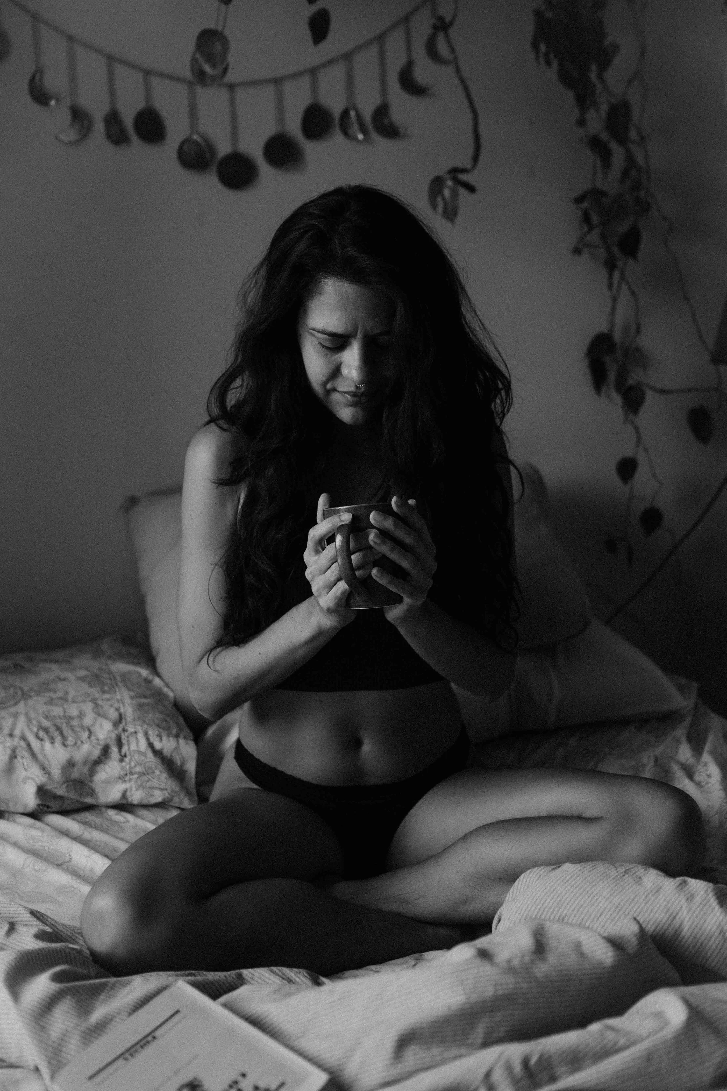 A woman with long dark hair sitting cross-legged on a bed, holding a mug and looking down at it thoughtfully in a dimly lit room.