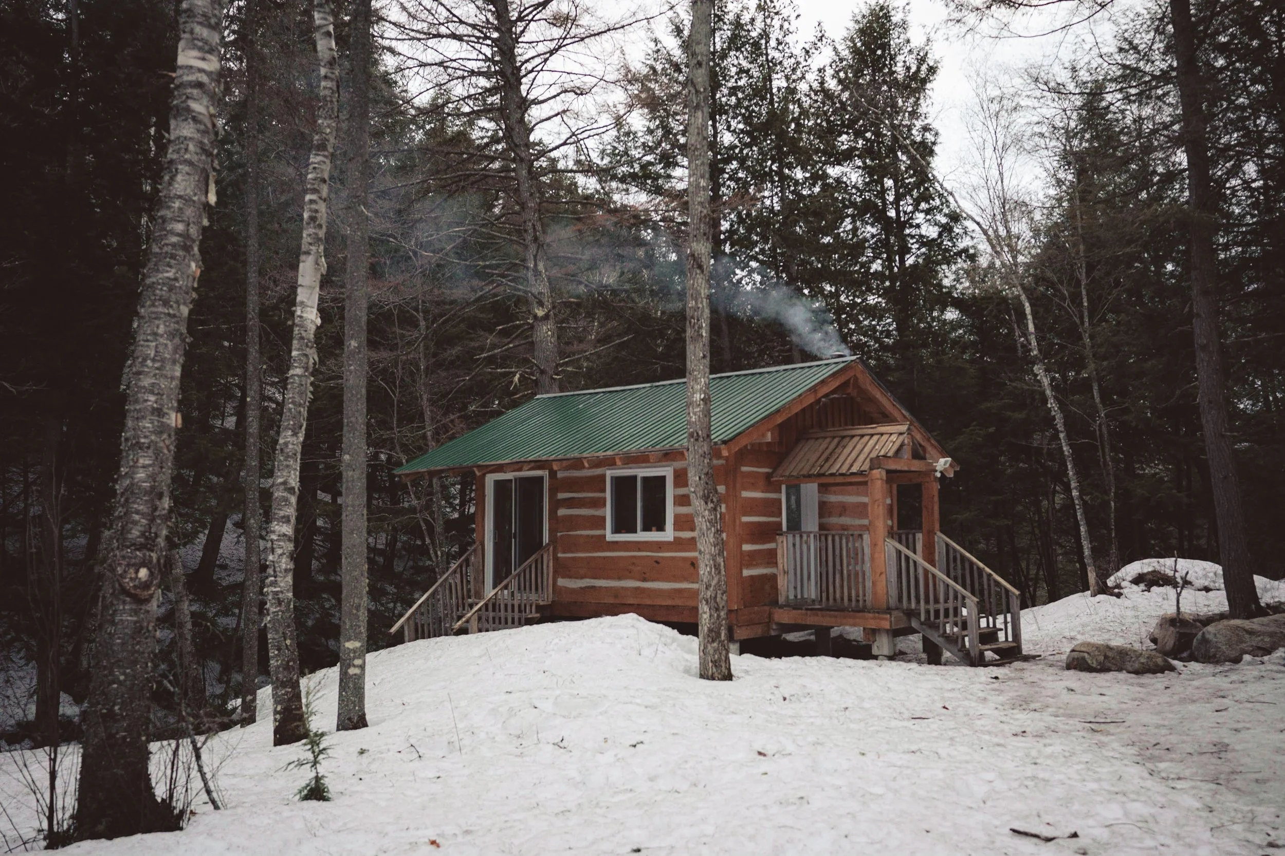 A small wooden cabin with a green metal roof, surrounded by snow-covered ground and tall trees in a forested area.