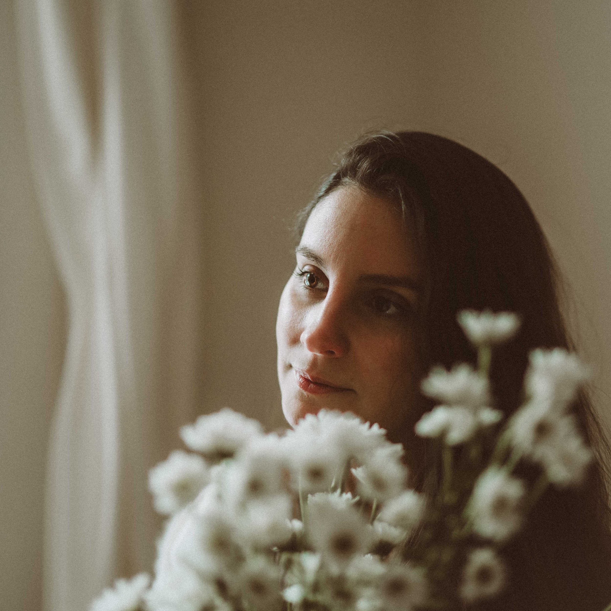 A woman gazing thoughtfully through a window with a bouquet of white flowers in front of her.