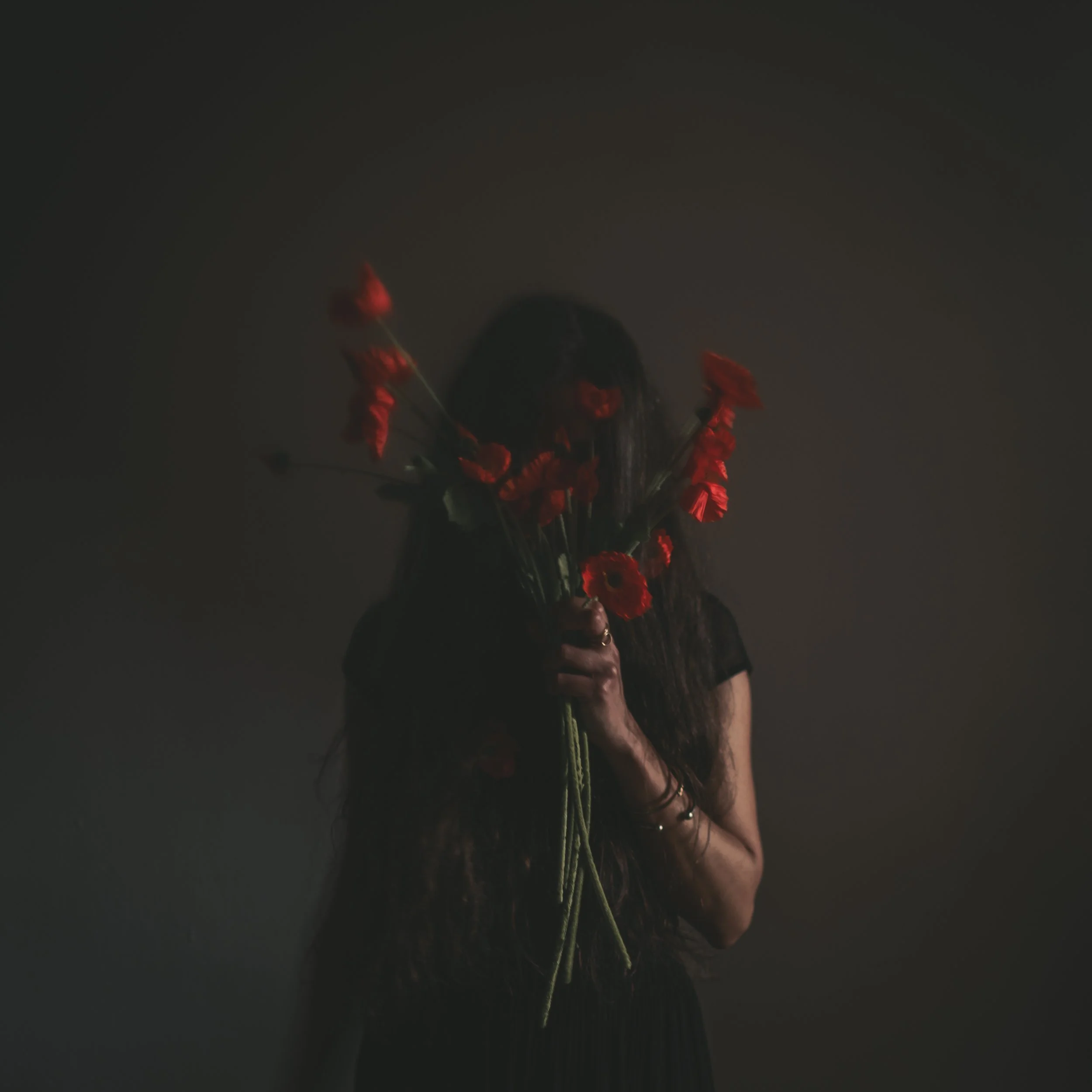 A woman with long dark hair holding a bouquet of red flowers in front of her face in a dimly lit setting.
