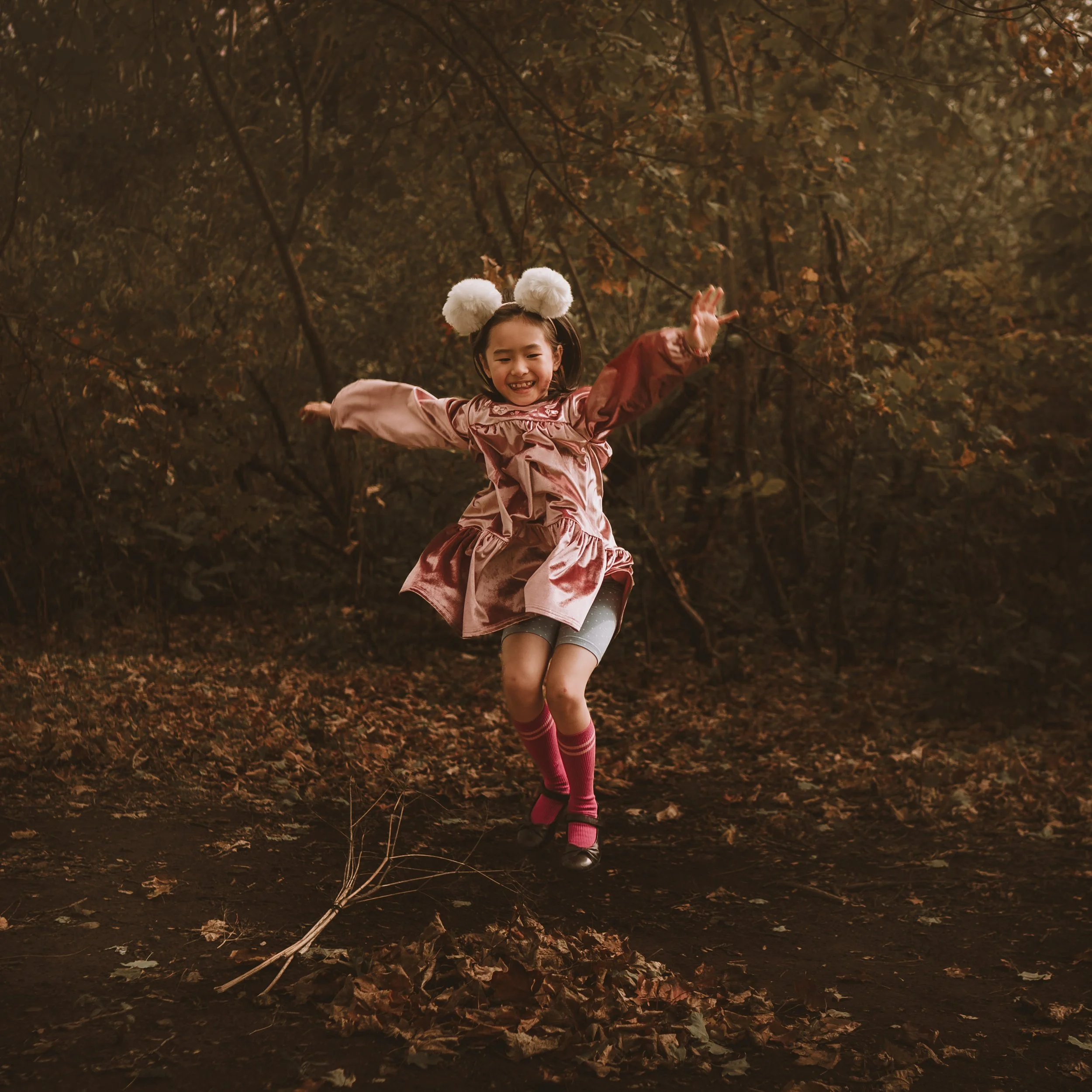 A girl wearing a pink dress, gray leggings, pink socks, and black shoes is smiling and playing on a swing in a wooded area with fallen leaves on the ground. She has white fluffy pom-pom headbands and long dark hair.