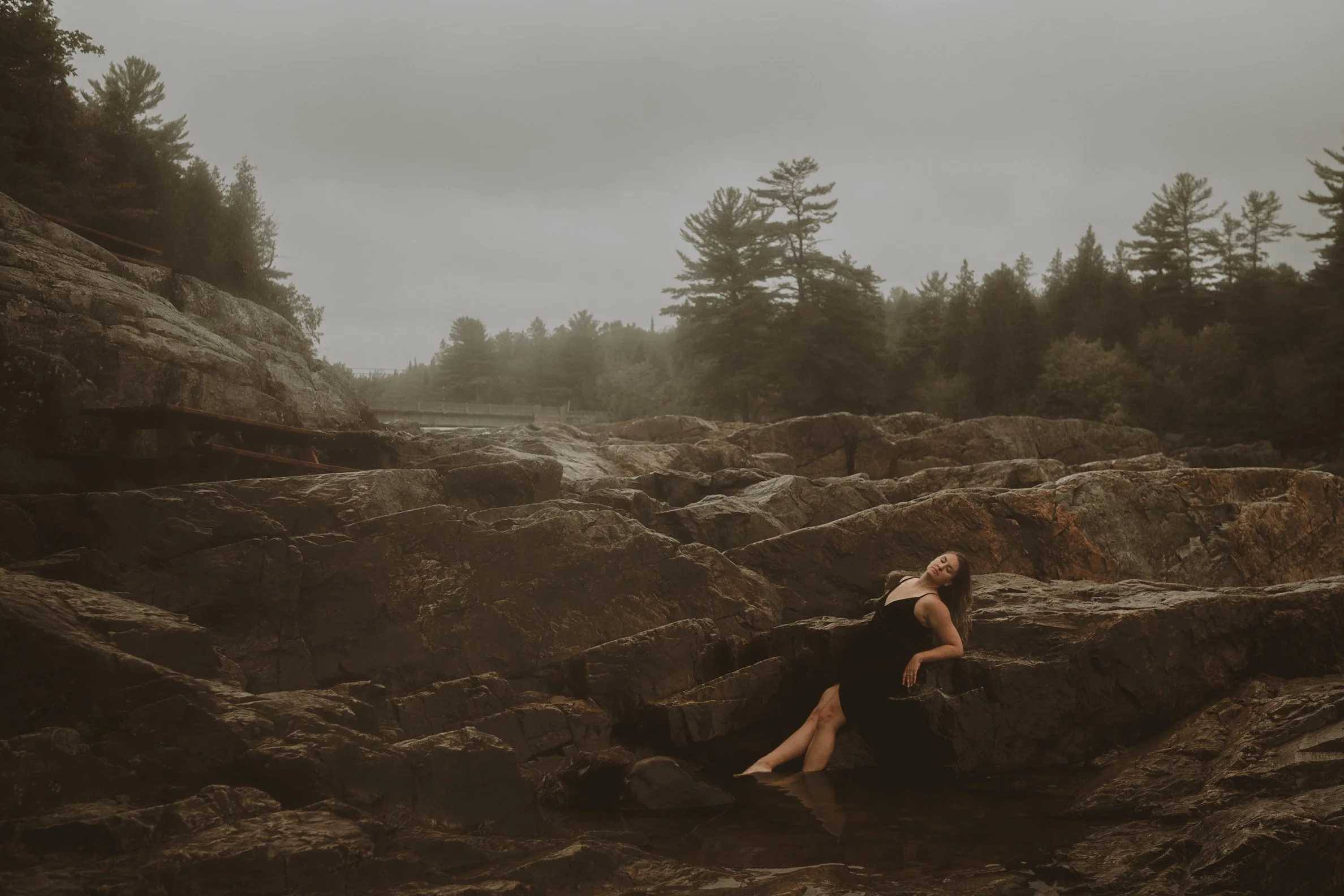 A woman in a black dress sitting on rocks in a natural outdoor setting with trees and cloudy sky in the background.