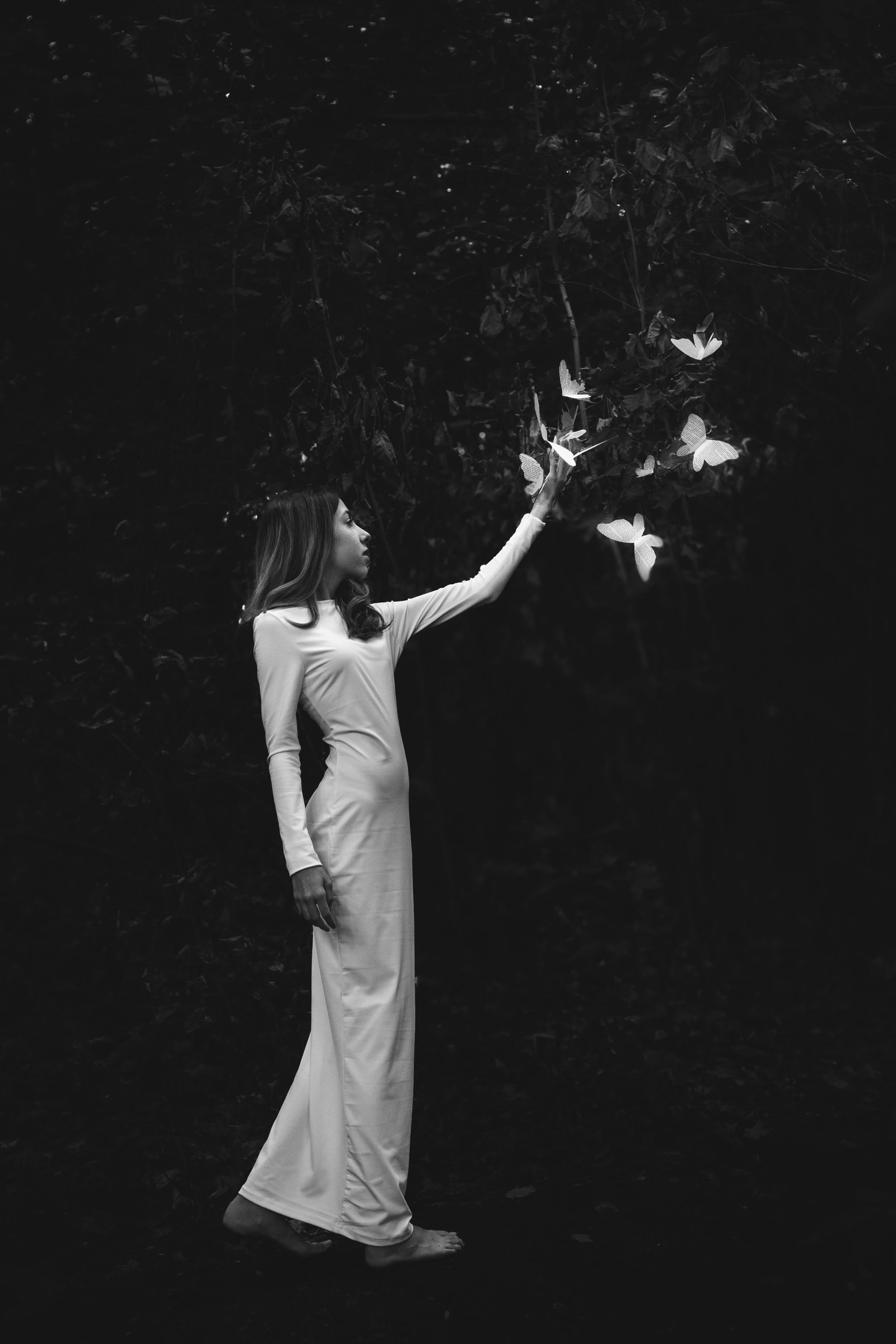 A woman in a white dress standing barefoot outdoors in a dark environment, reaching out to sky to release white butterflies.