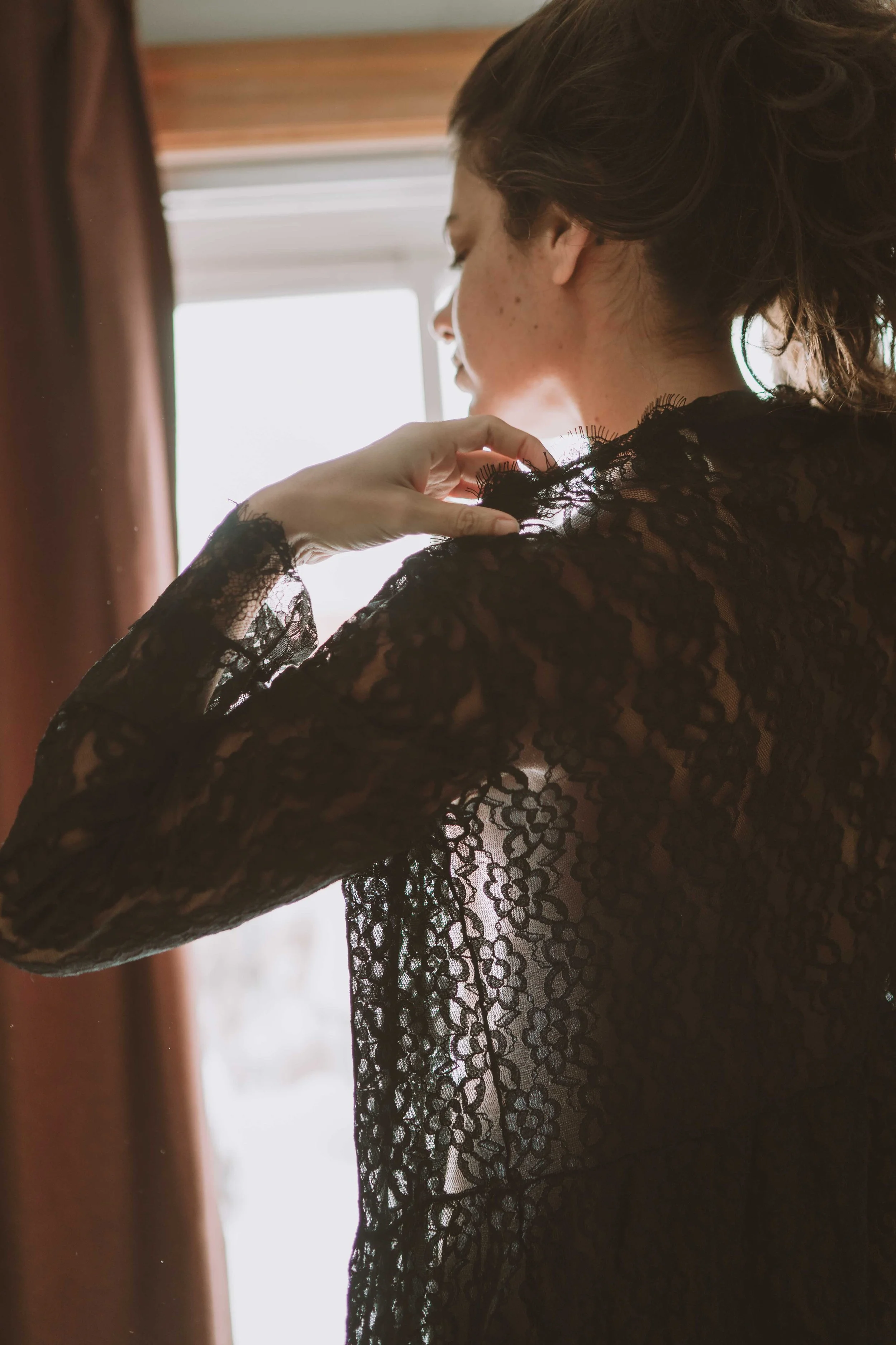 A woman with light skin and wavy brown hair wearing a black lace top, adjusting her lace sleeve near a window with curtains in the background.