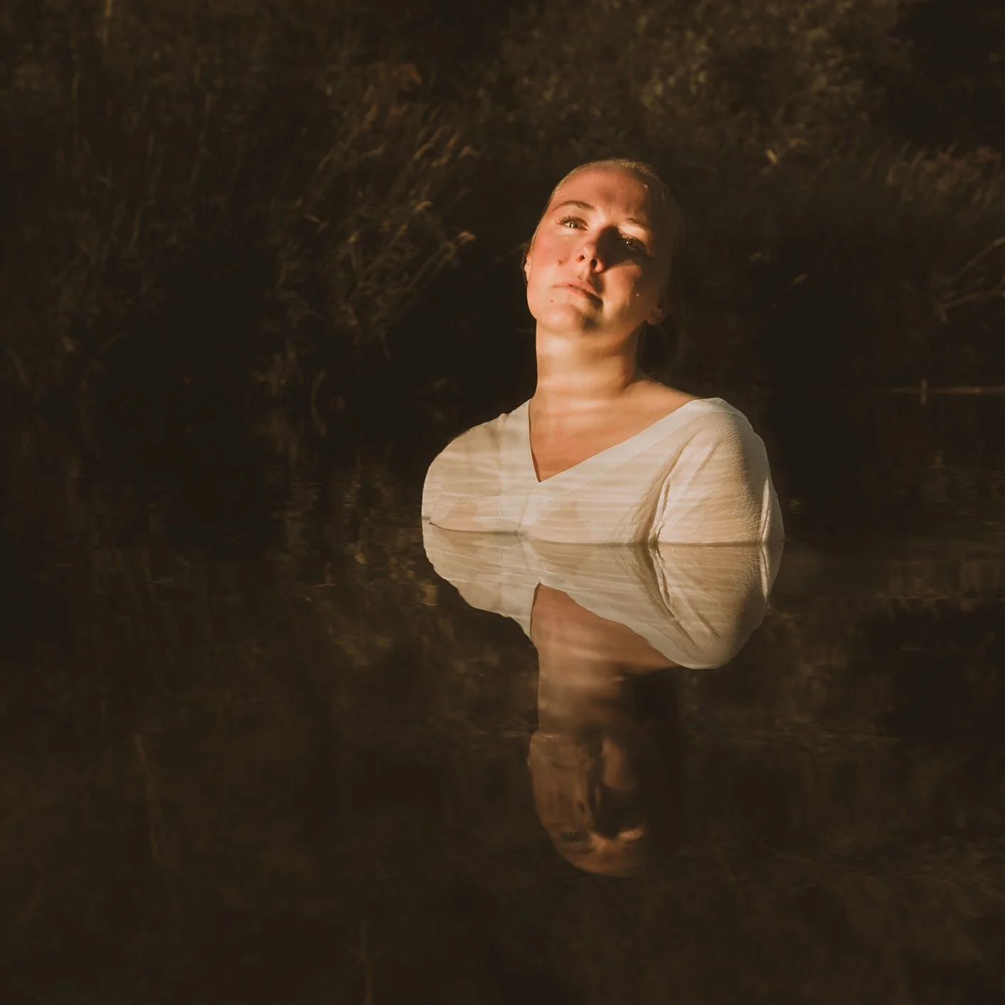 Woman with light skin and reddish-brown hair, wearing a white shirt, standing in water with warm lighting, dark background