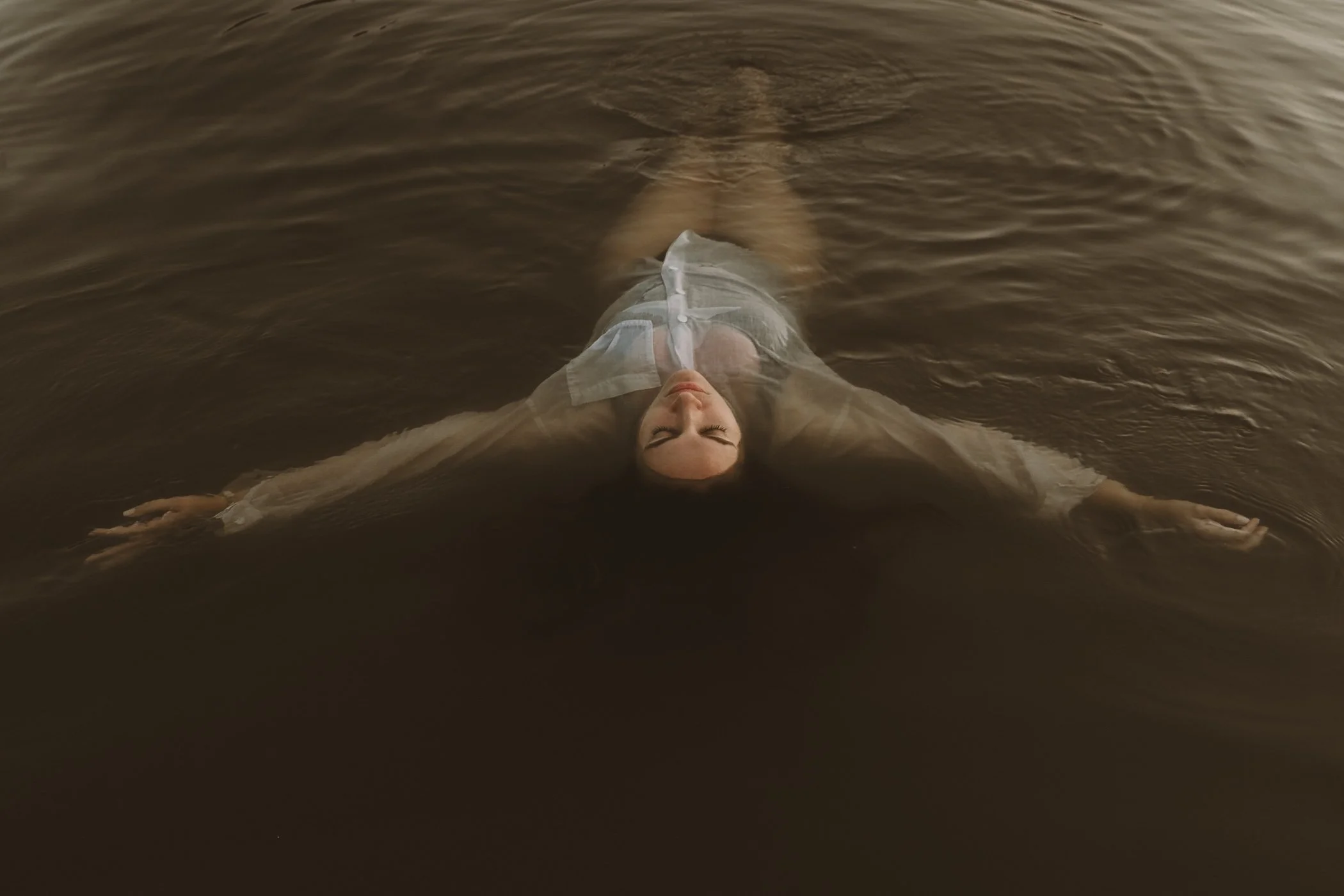 A person lies back in shallow, dark water with eyes closed, wearing a sheer, long-sleeved dress, with arms outstretched, face calm, and water rippling around them.
