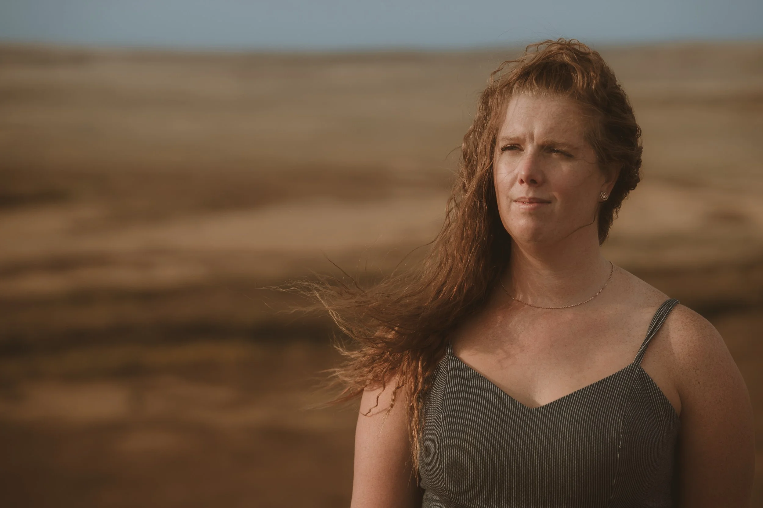 A woman with long curly red hair in a striped gray dress standing on a beach with wind blowing her hair, looking into the distance.