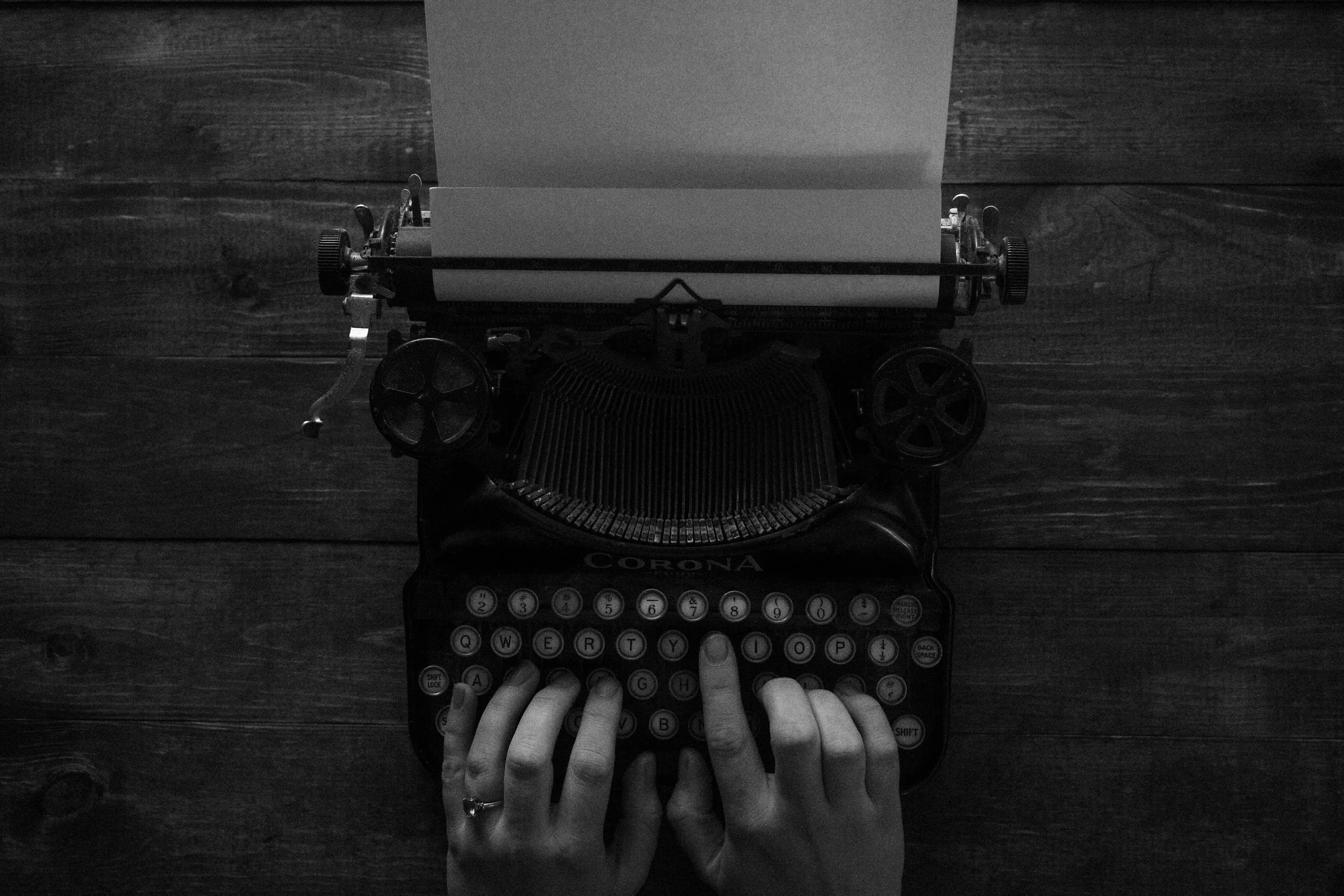 A black and white grainy image of two hands resting on the keys of an old fashioned typewriter with a black piece of paper loaded.