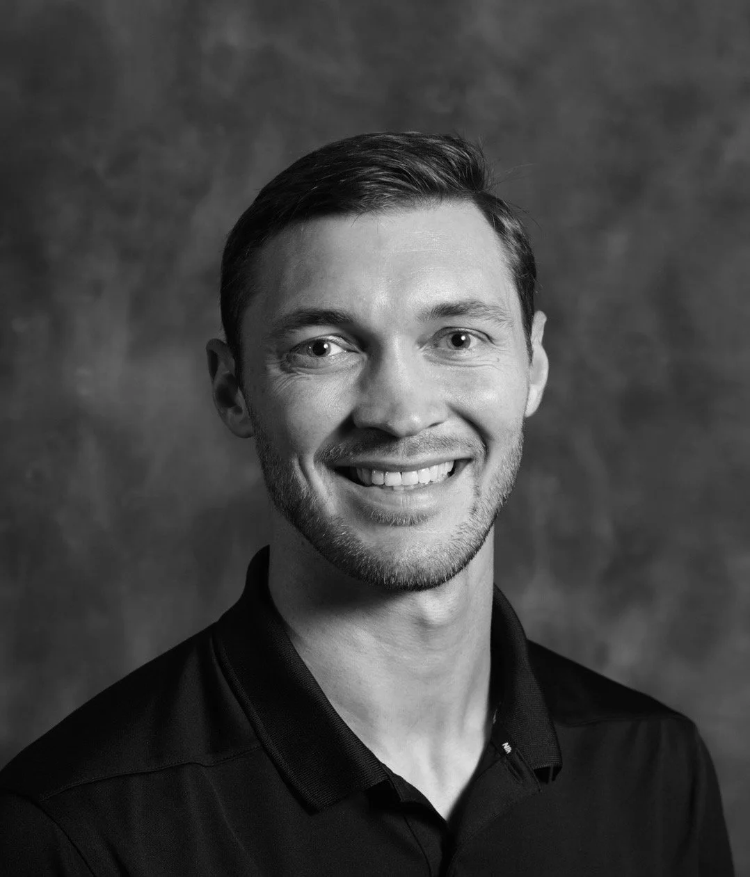 Black and white portrait of a smiling young man with short dark hair, wearing a collared shirt, against a textured background.