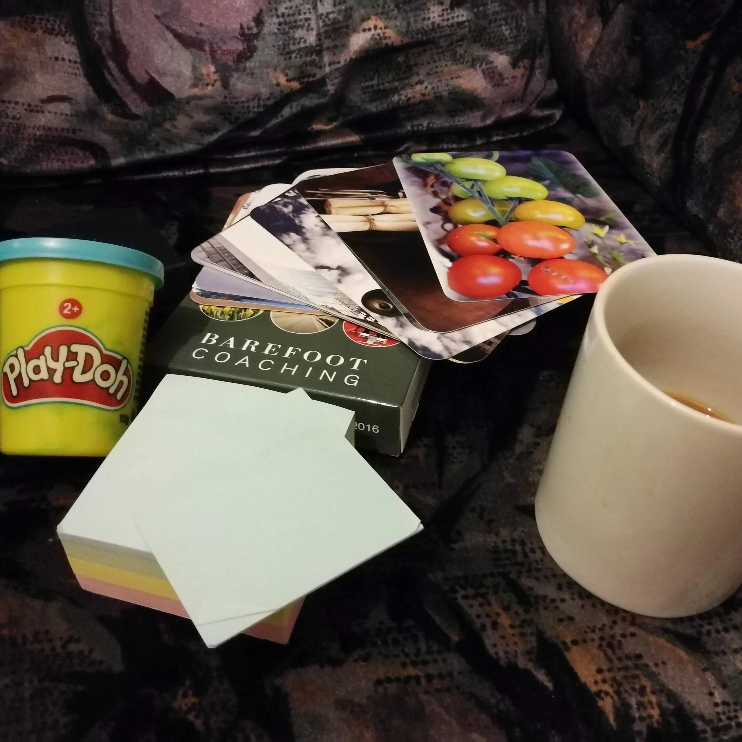 A Coach's Tools on a flowery sofa: Play-doh in a yellow pot, with blue lid, a stack of picture cards with red-green tomatoes uppermost,  a pile of post-it notes, one peeled back and a white mug with most of a drink consumed on the right